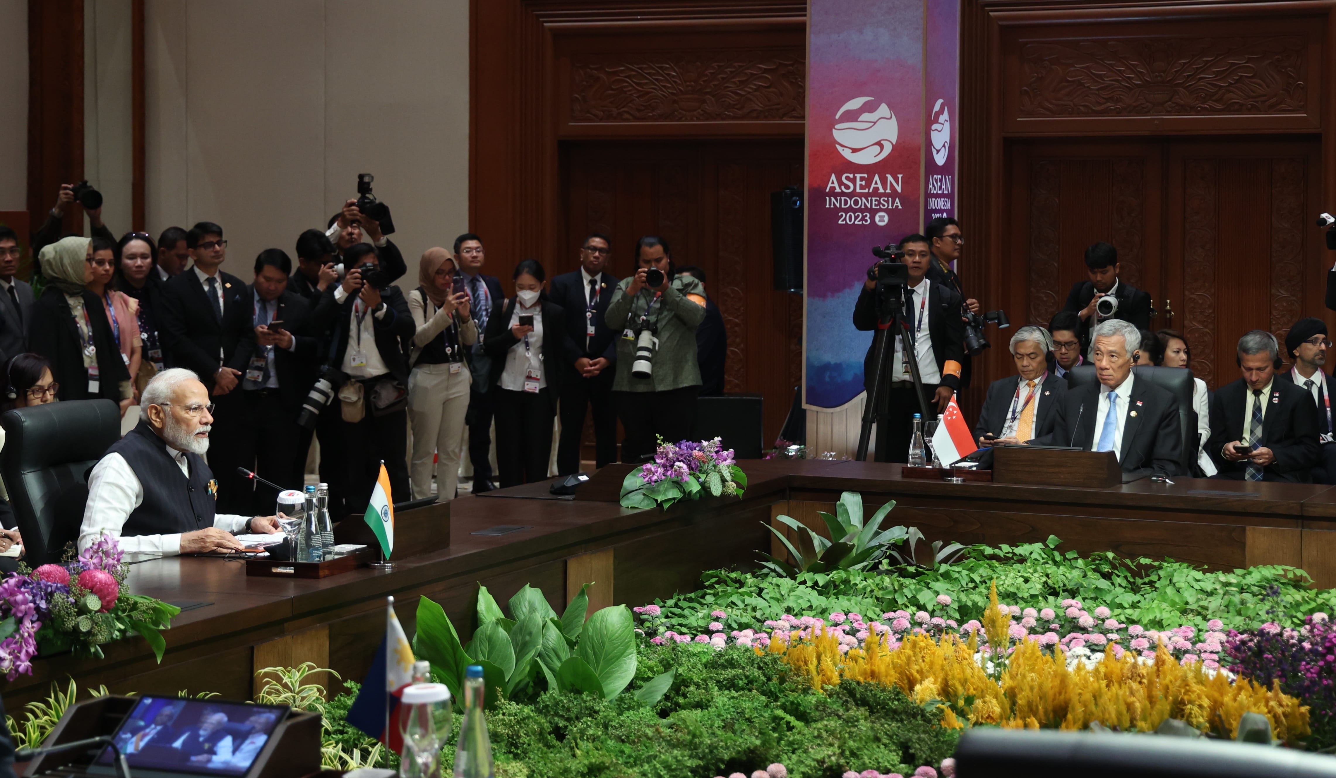 ASEAN summit with dignitaries at tables, floral display, and many press photographers.