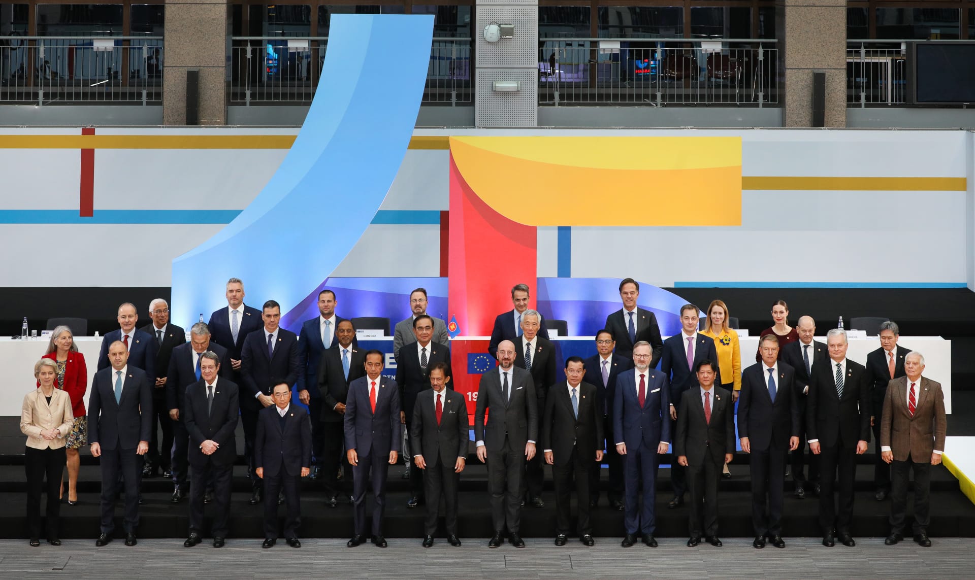 Group poses in front of a backdrop with blue, red, and yellow blocks. Most wear suits.