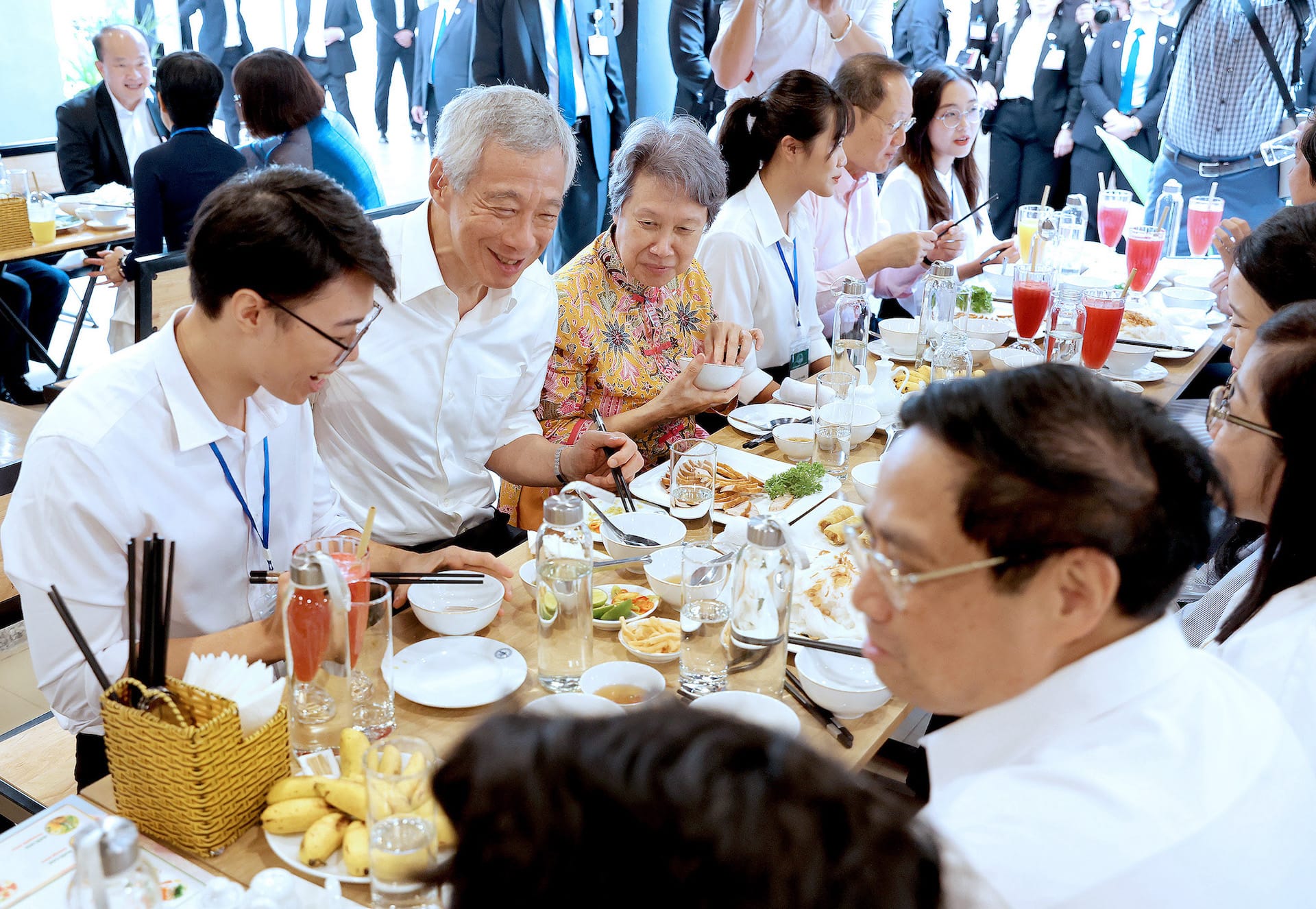 Group eating at a table with various dishes, drinks, and utensils. Lee Hsien Loong visible.