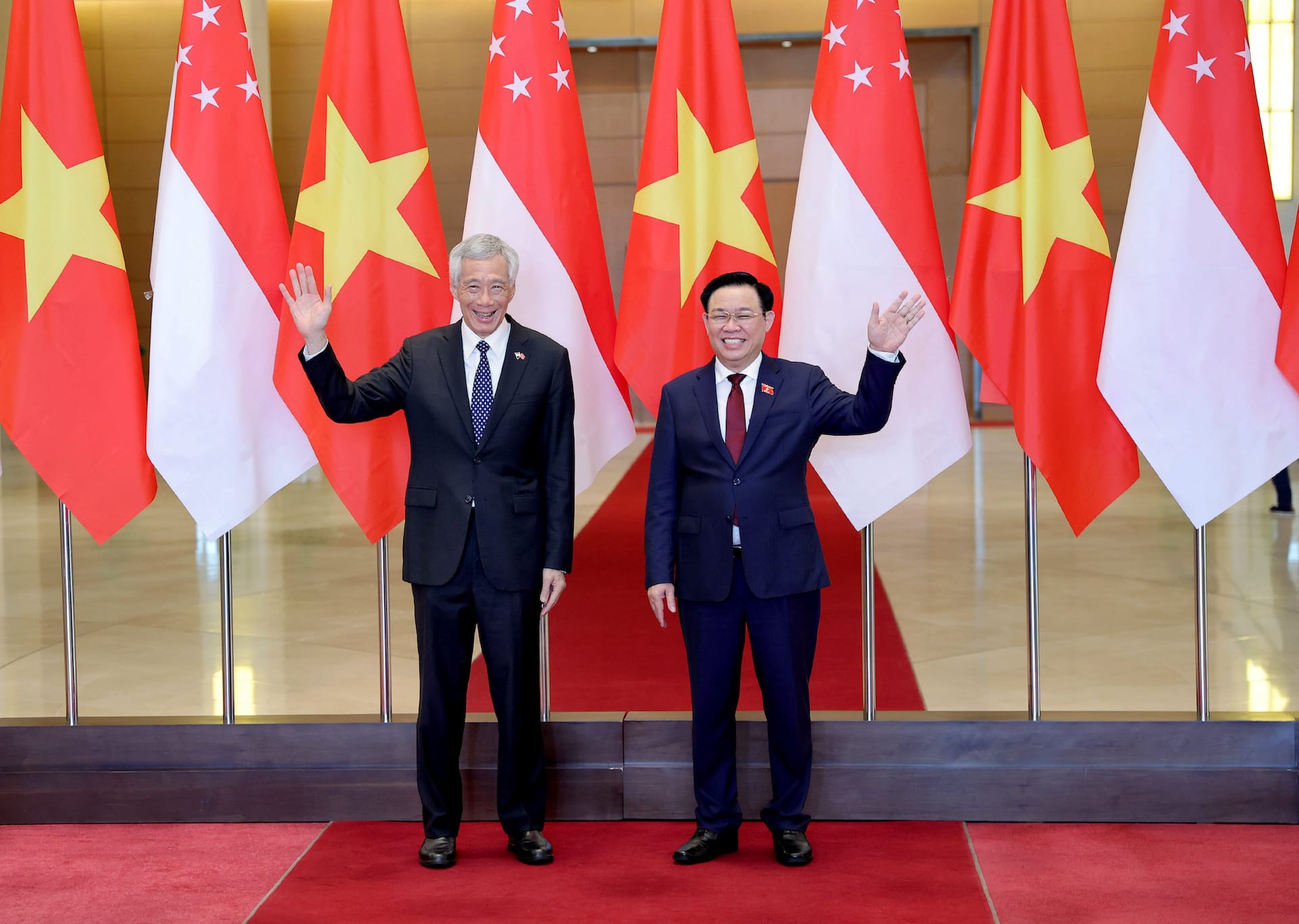 Two men in suits waving, standing before Singapore and Vietnam flags.