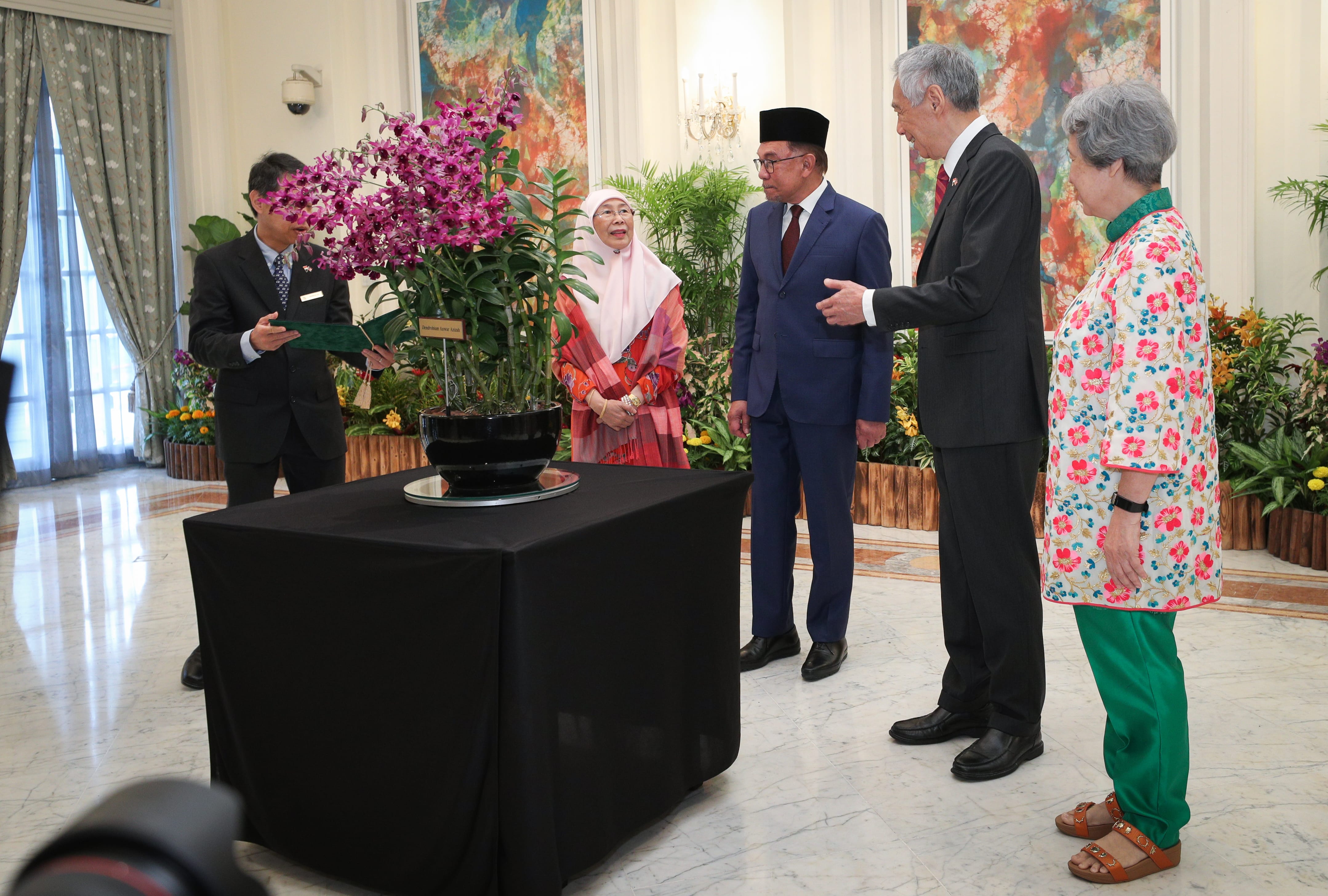 Dignitaries and staff standing near orchid arrangement with "Dendrobium Anwar Azizah" label.