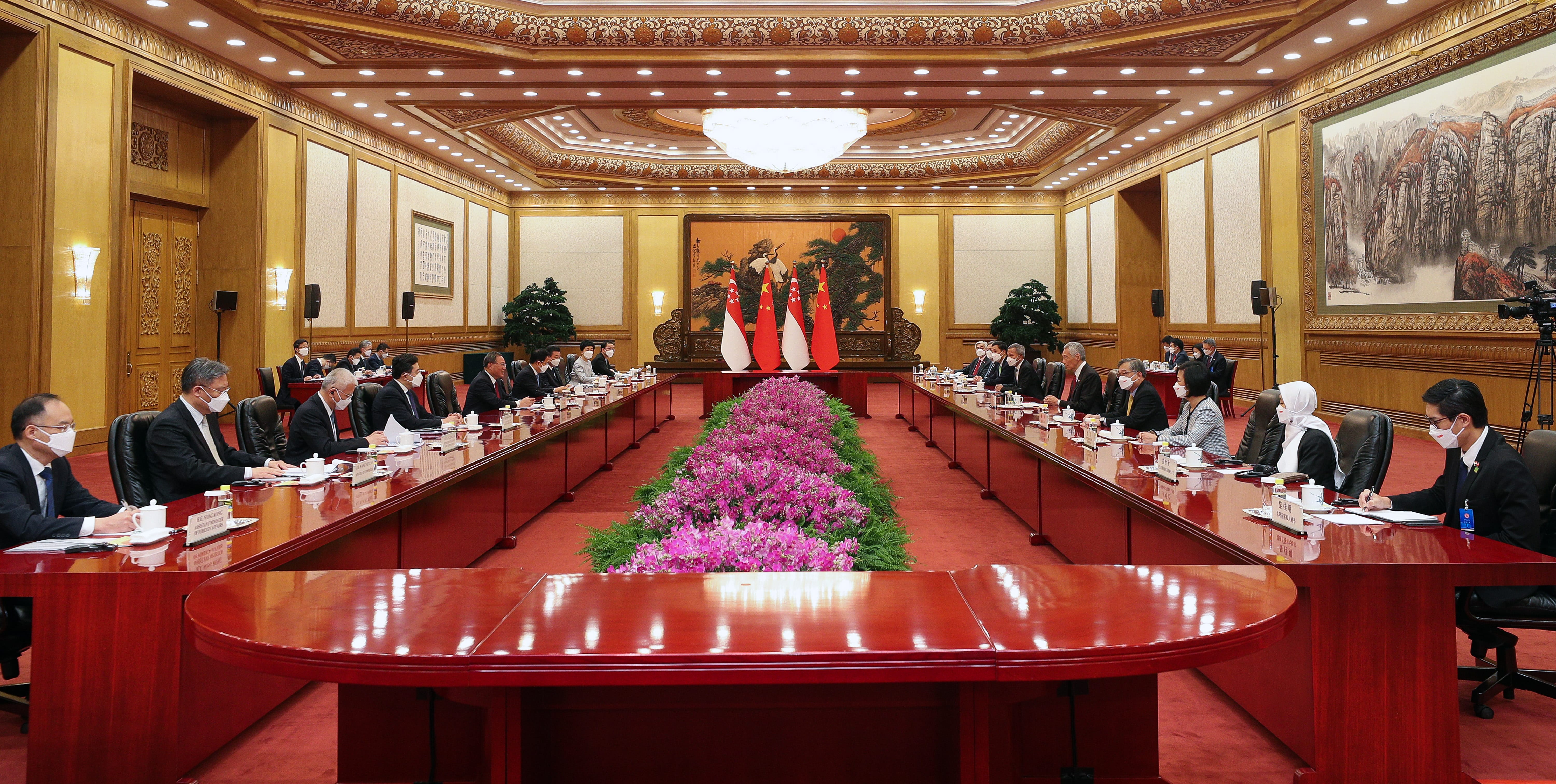 People sit around long red conference table with flags and flowers in ornate hall.