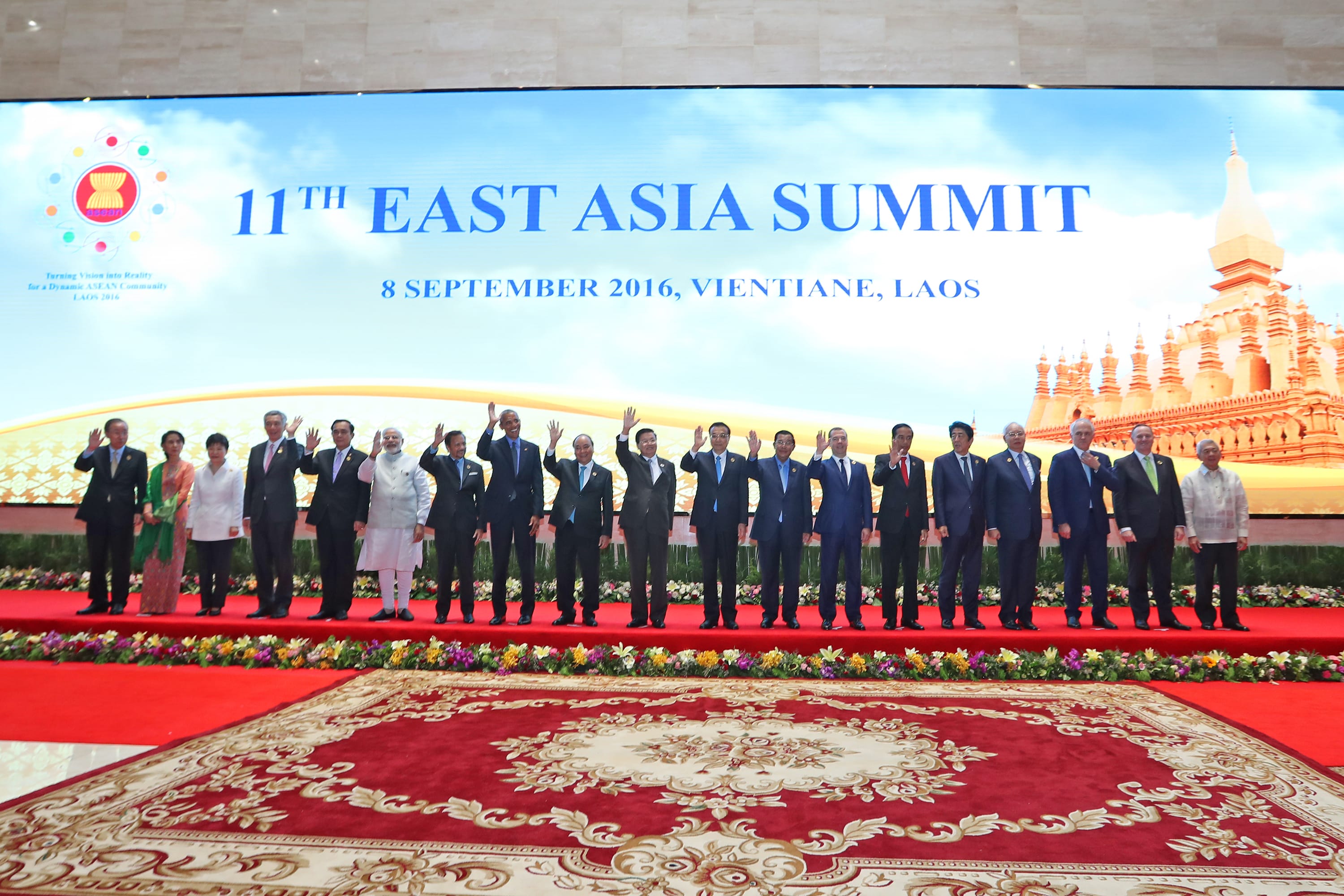 Group of people on stage in front of "11th East Asia Summit" backdrop, waving.