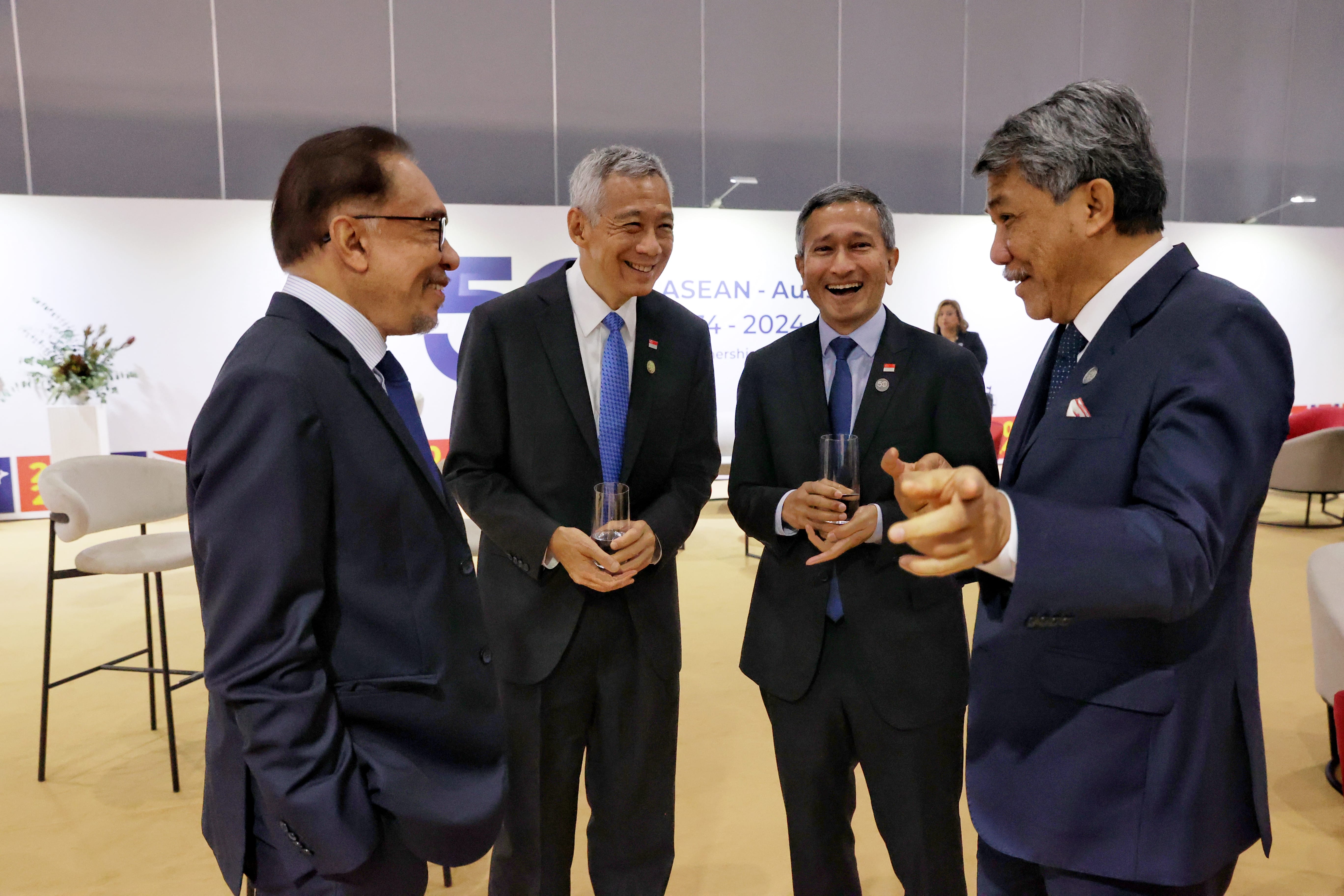 Four men in suits talking, holding glasses; ASEAN backdrop.