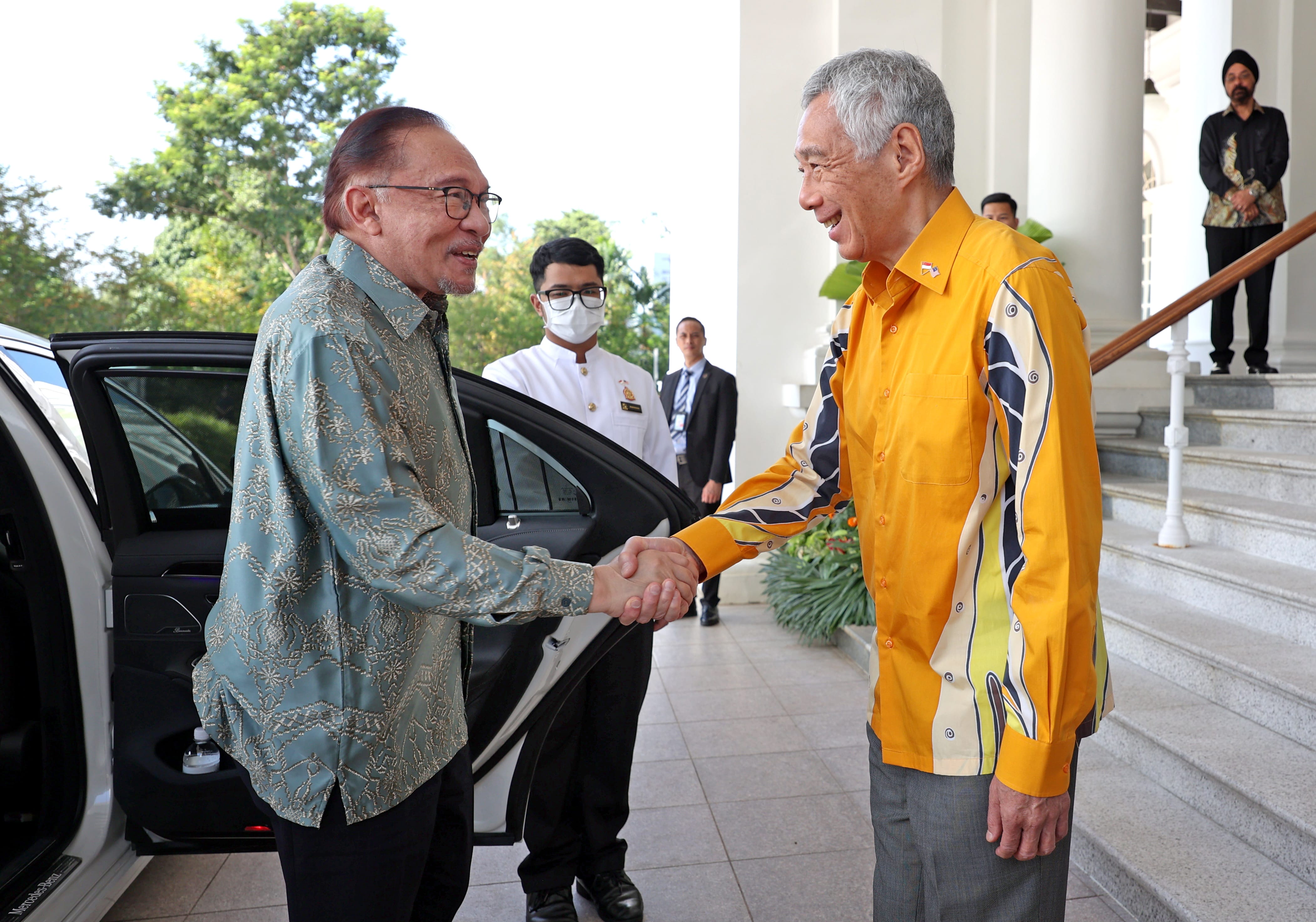 Anwar Ibrahim and Lee Hsien Loong shake hands in front of a Mercedes, several people stand behind them.