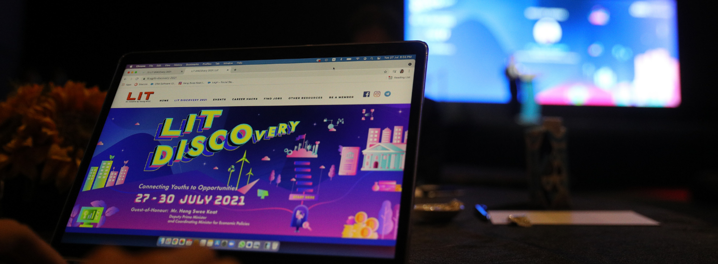 Laptop screen shows LIT DISCOvery website with cityscape design, a larger monitor blurs in the background.
