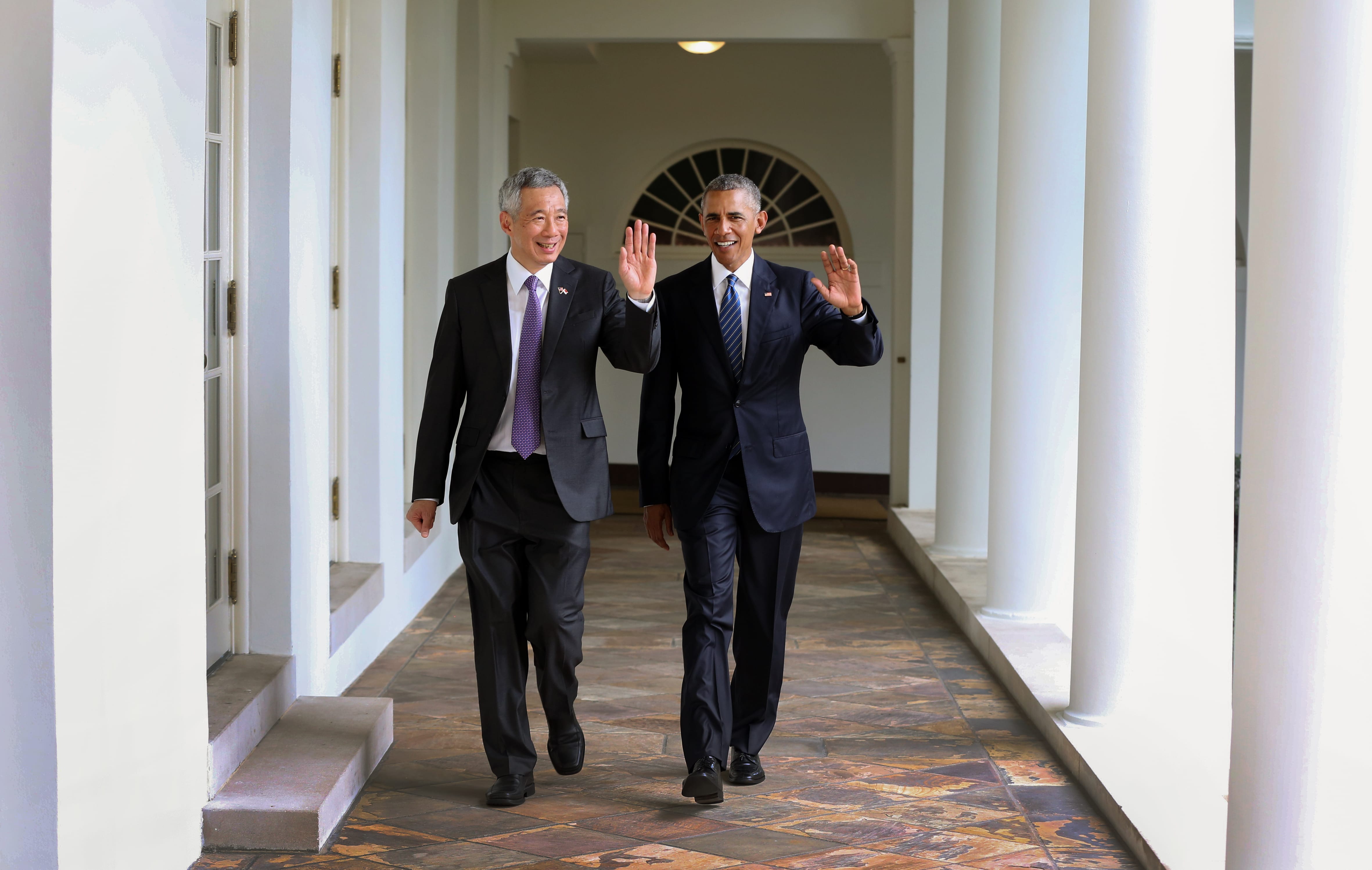 Obama and Lee Hsien Loong waving, walking along a colonnade.