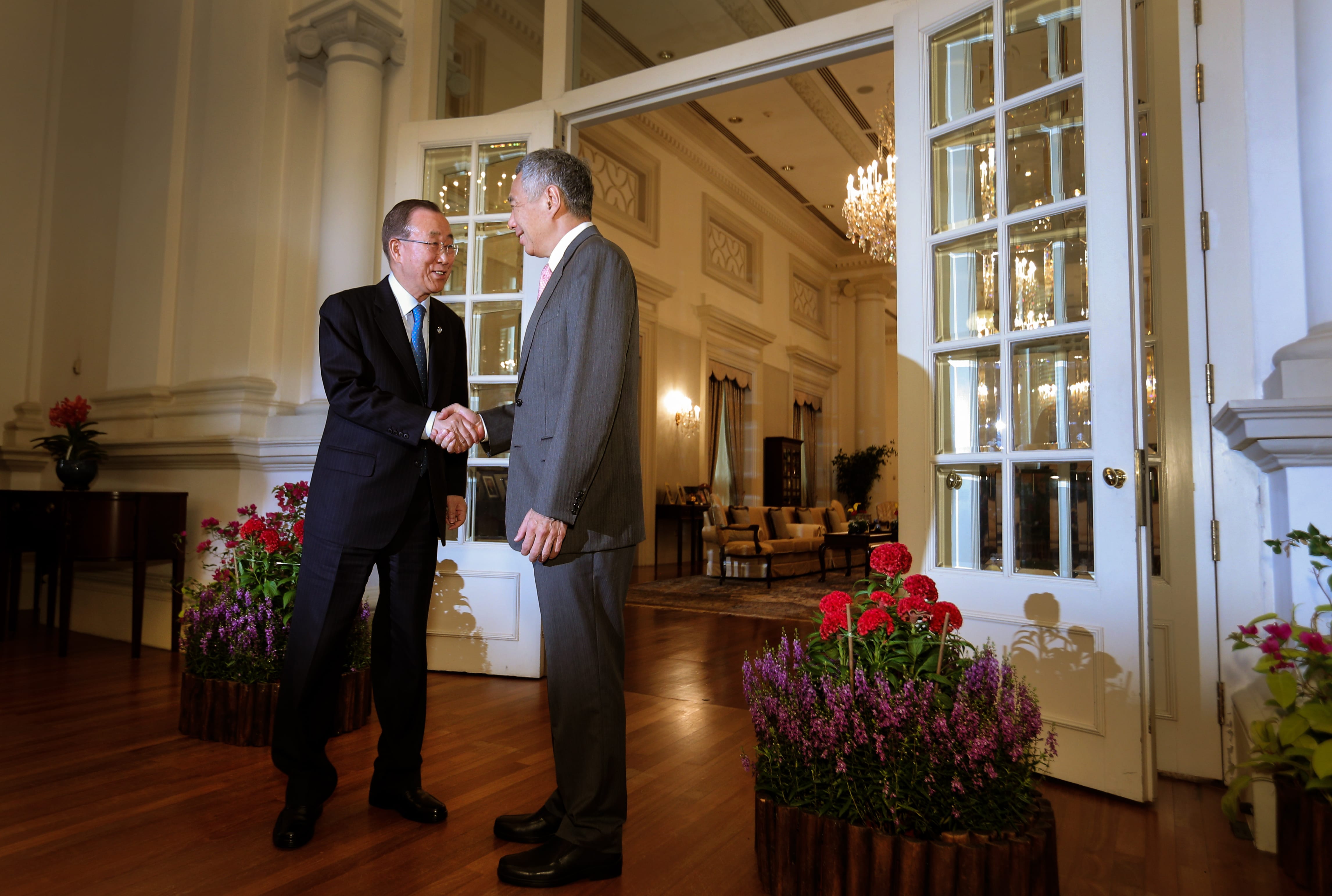 Ban Ki-moon shaking hands with a man in suits near white double doors. Interior view with chandelier.