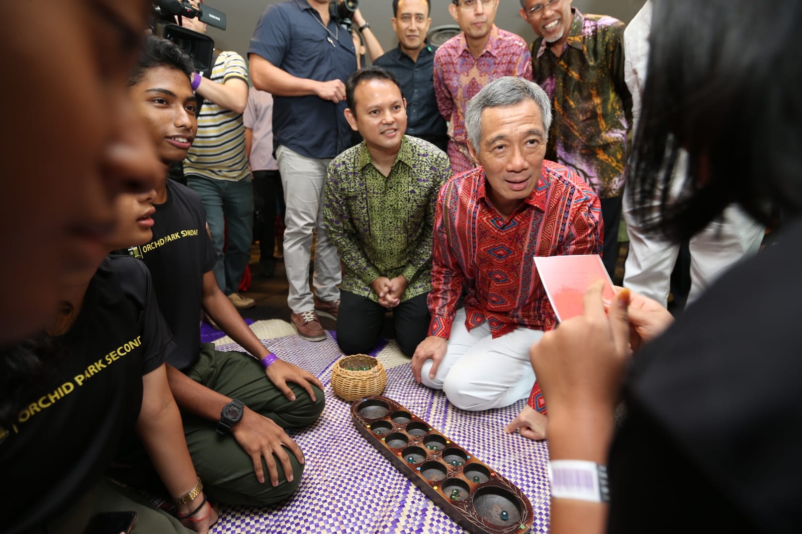Lee Hsien Loong playing Congkak on a mat with people watching.