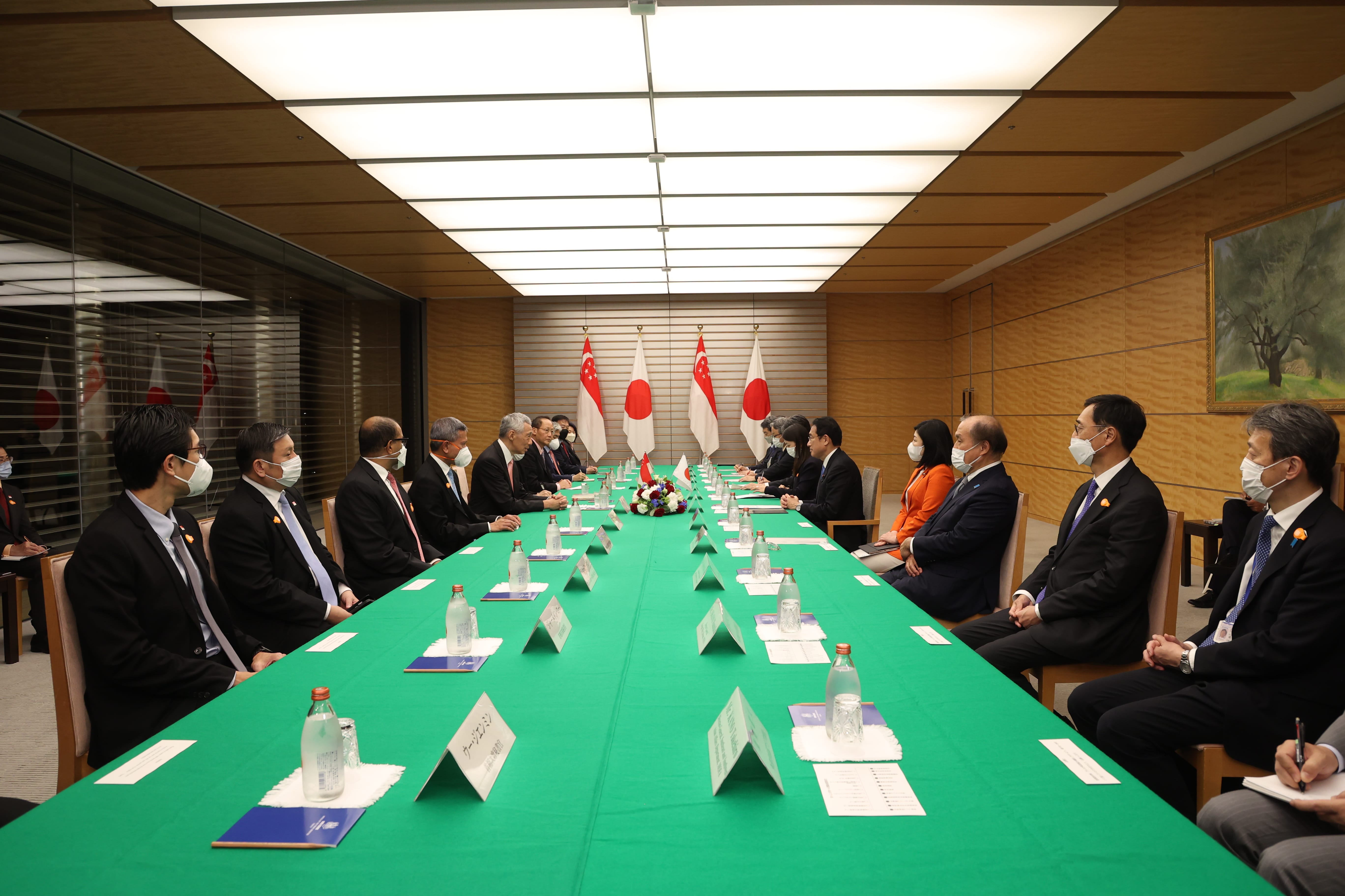 People in suits wearing face masks seated at long green table with Japanese and Singaporean flags.