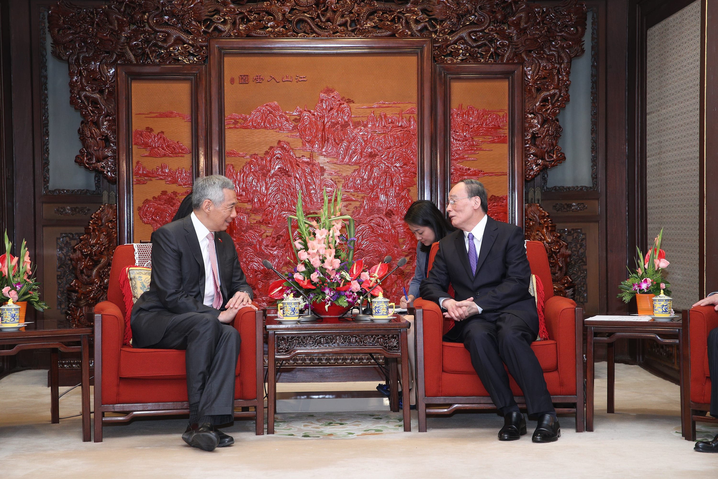 Two men in suits sit in red chairs facing each other in front of a large carved wooden backdrop.