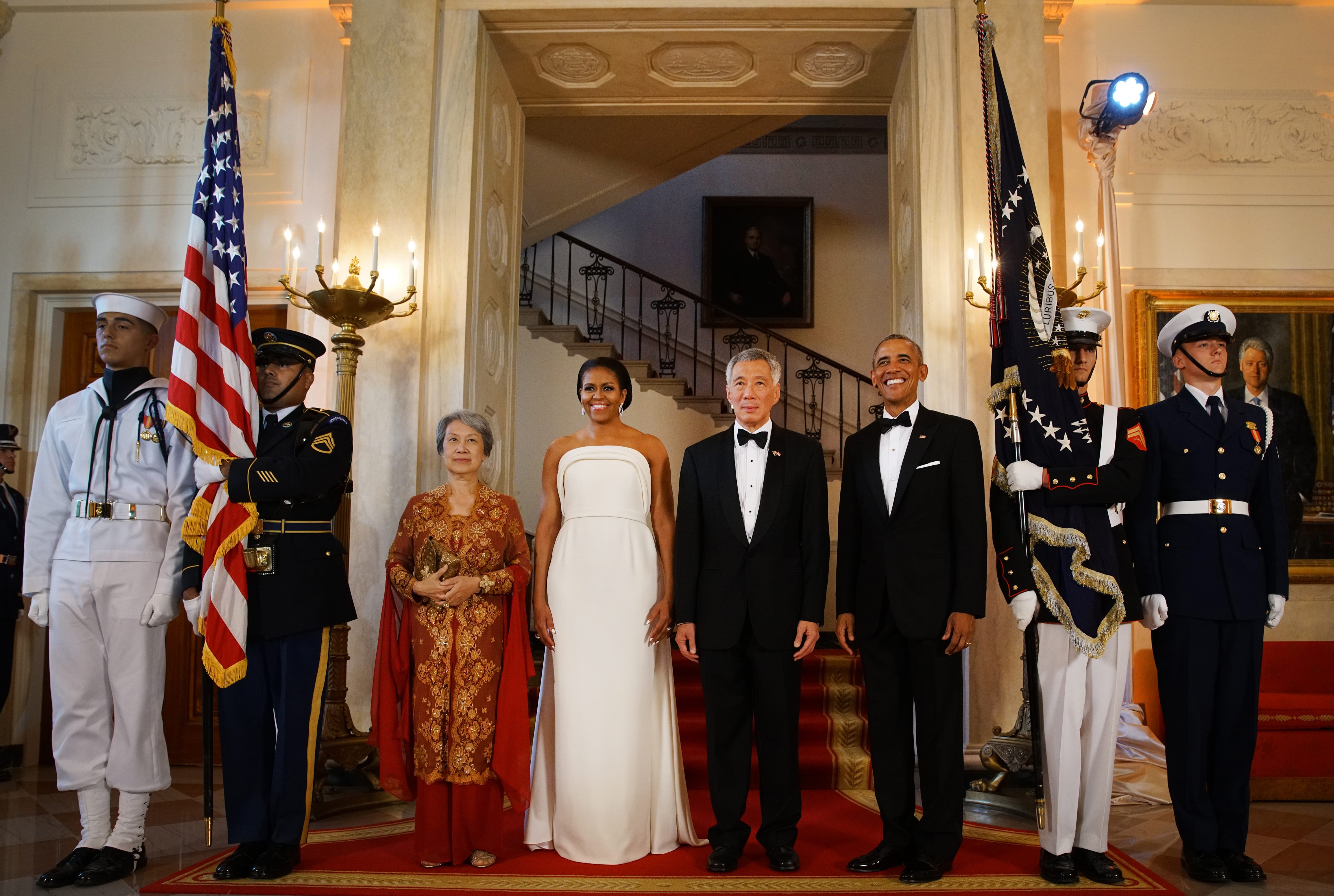 Obama family with Lee Hsien Loong and guards holding US flags, formal wear, ornate room.