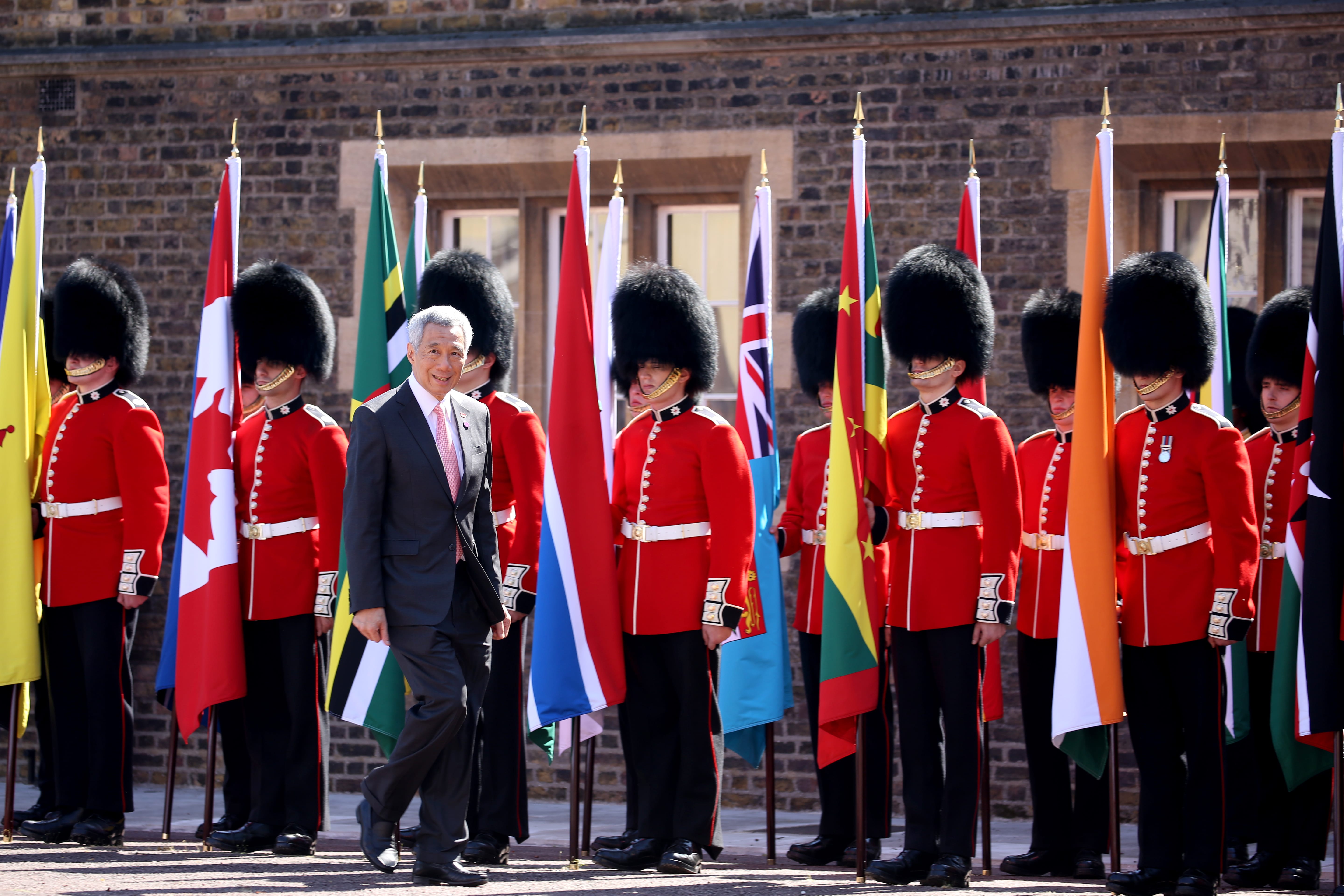 Lee Hsien Loong walks past guards in red coats holding flags against brick wall.