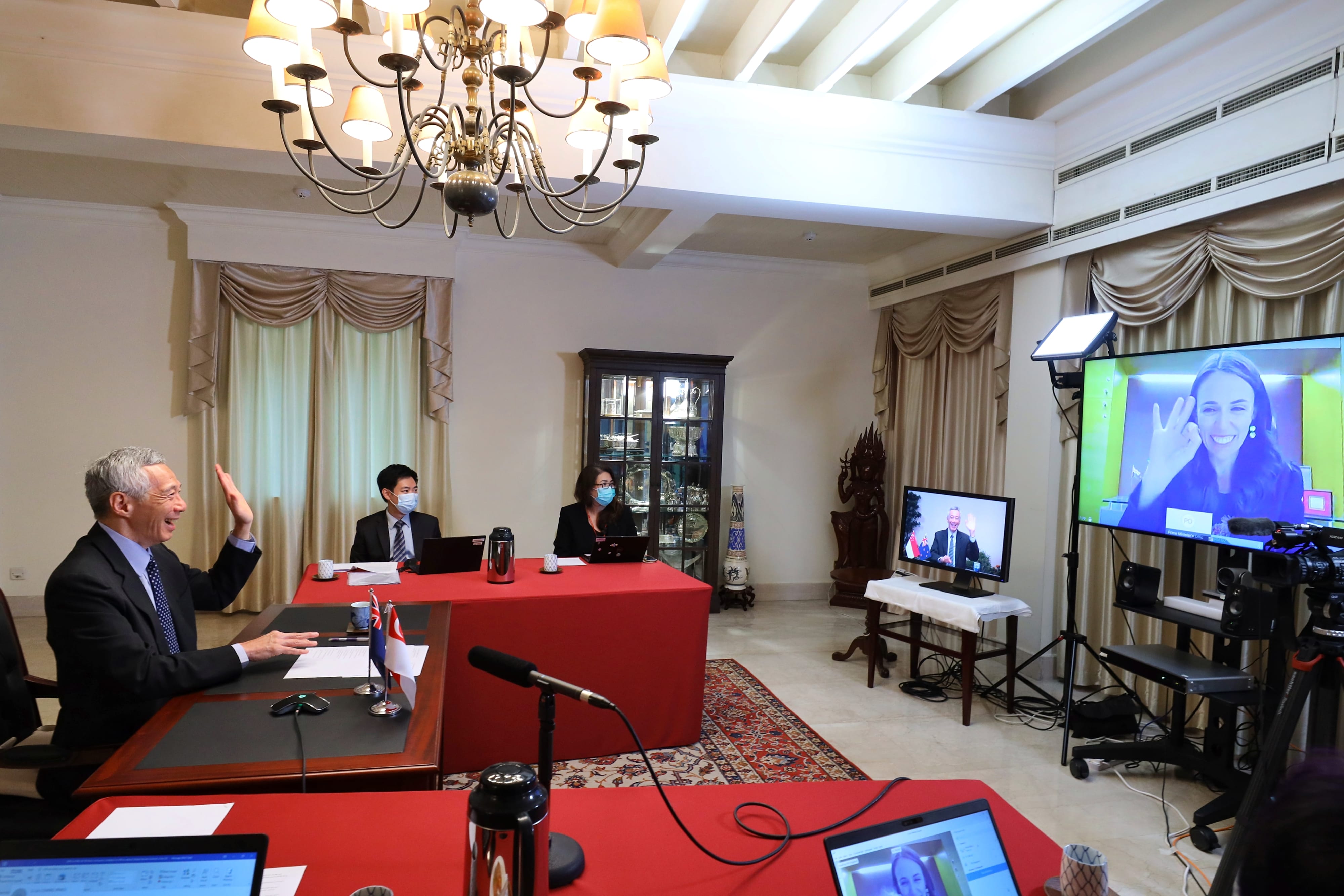Virtual meeting setup: People at desks with laptops, video displays of Lee Hsien Loong and Jacinda Ardern.