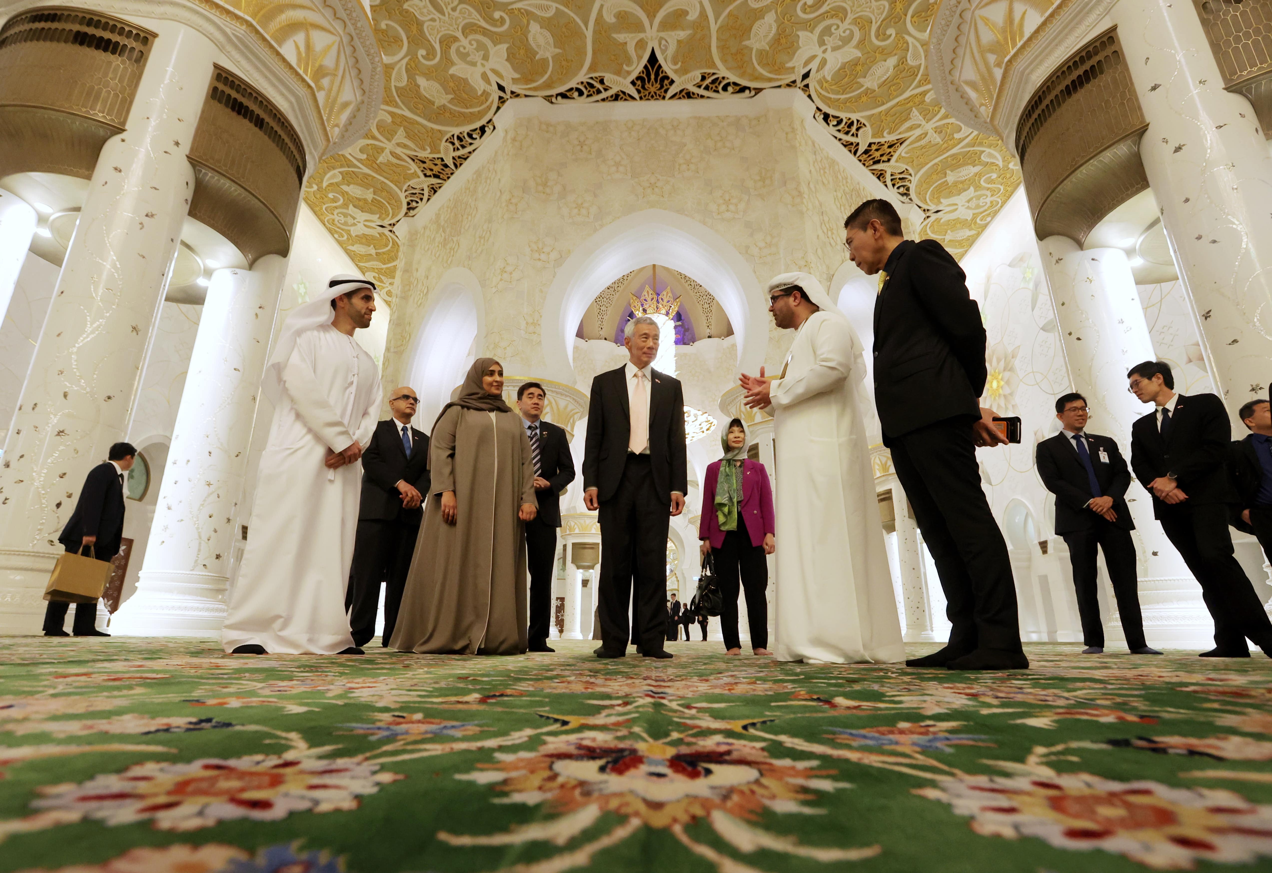 People in formal wear inside Sheikh Zayed Mosque. Ornate white walls and green carpet.