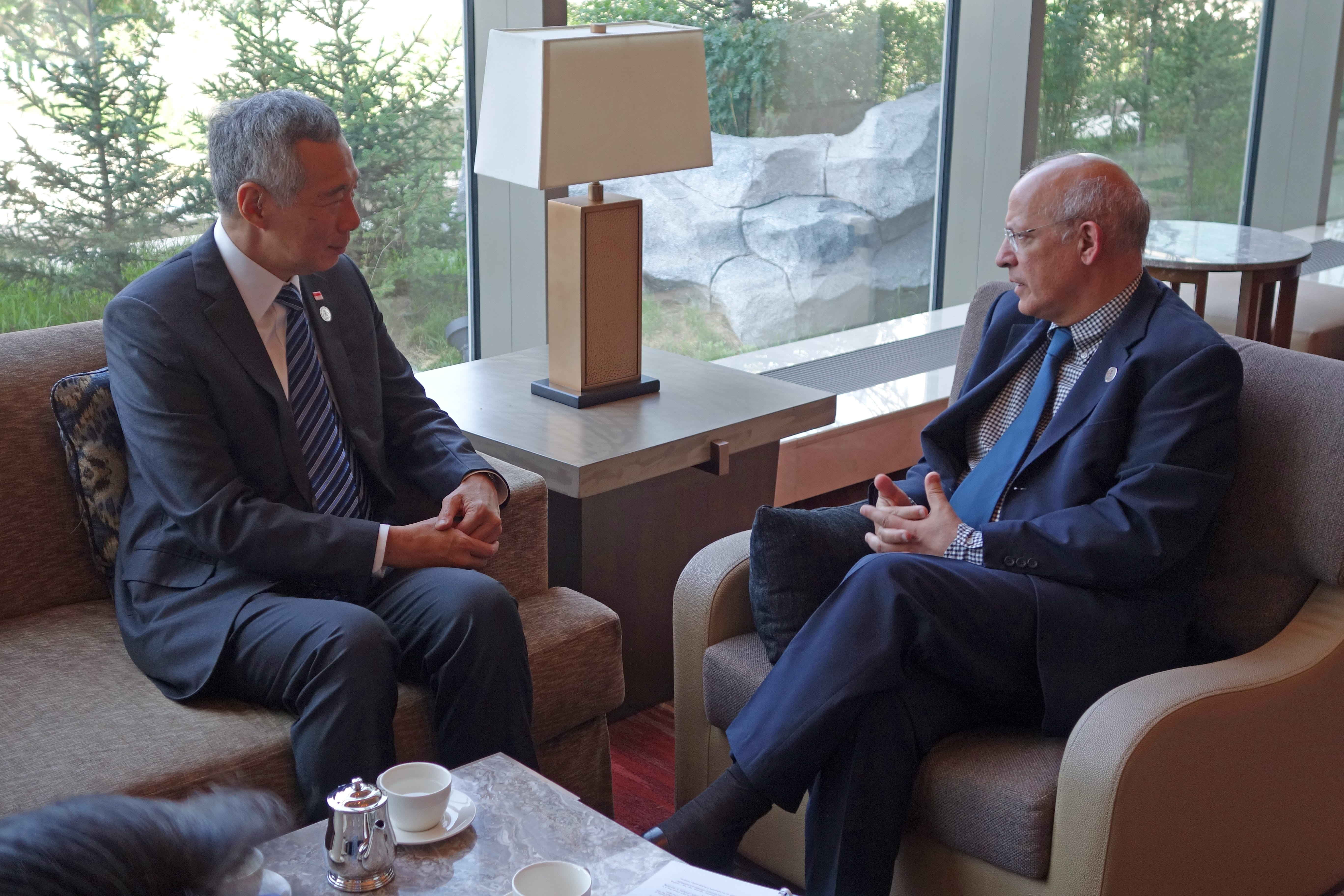 Two men in suits sit in armchairs, facing each other. Singapore flag visible on lapel. Tea service on a table.
