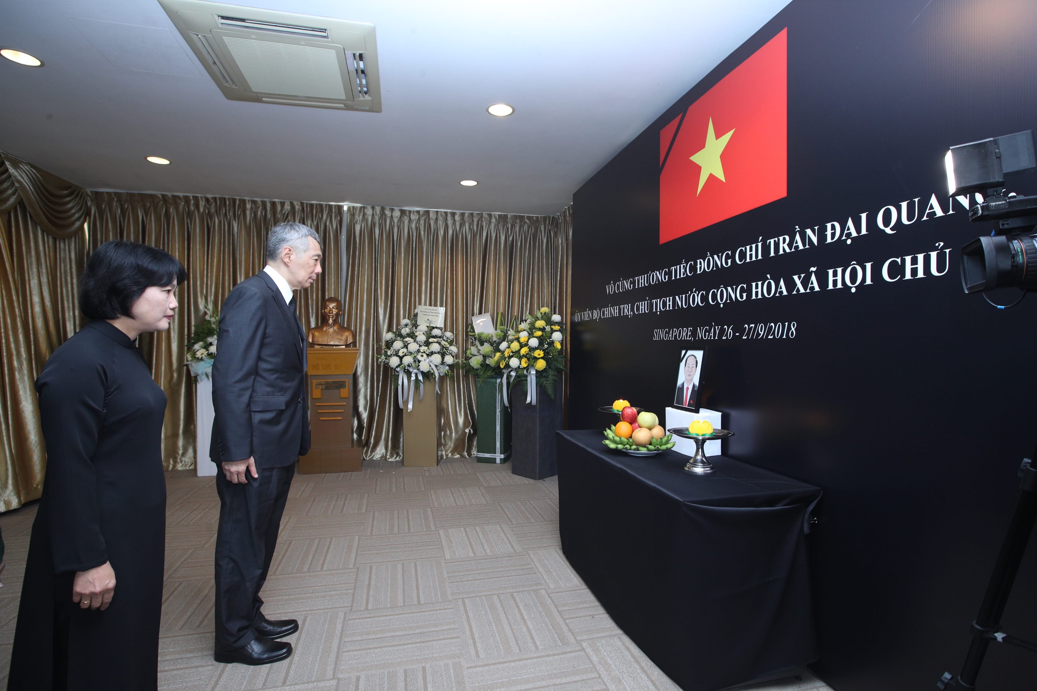 People stand near a Vietnamese flag, black backdrop with text, and a table with fruit.