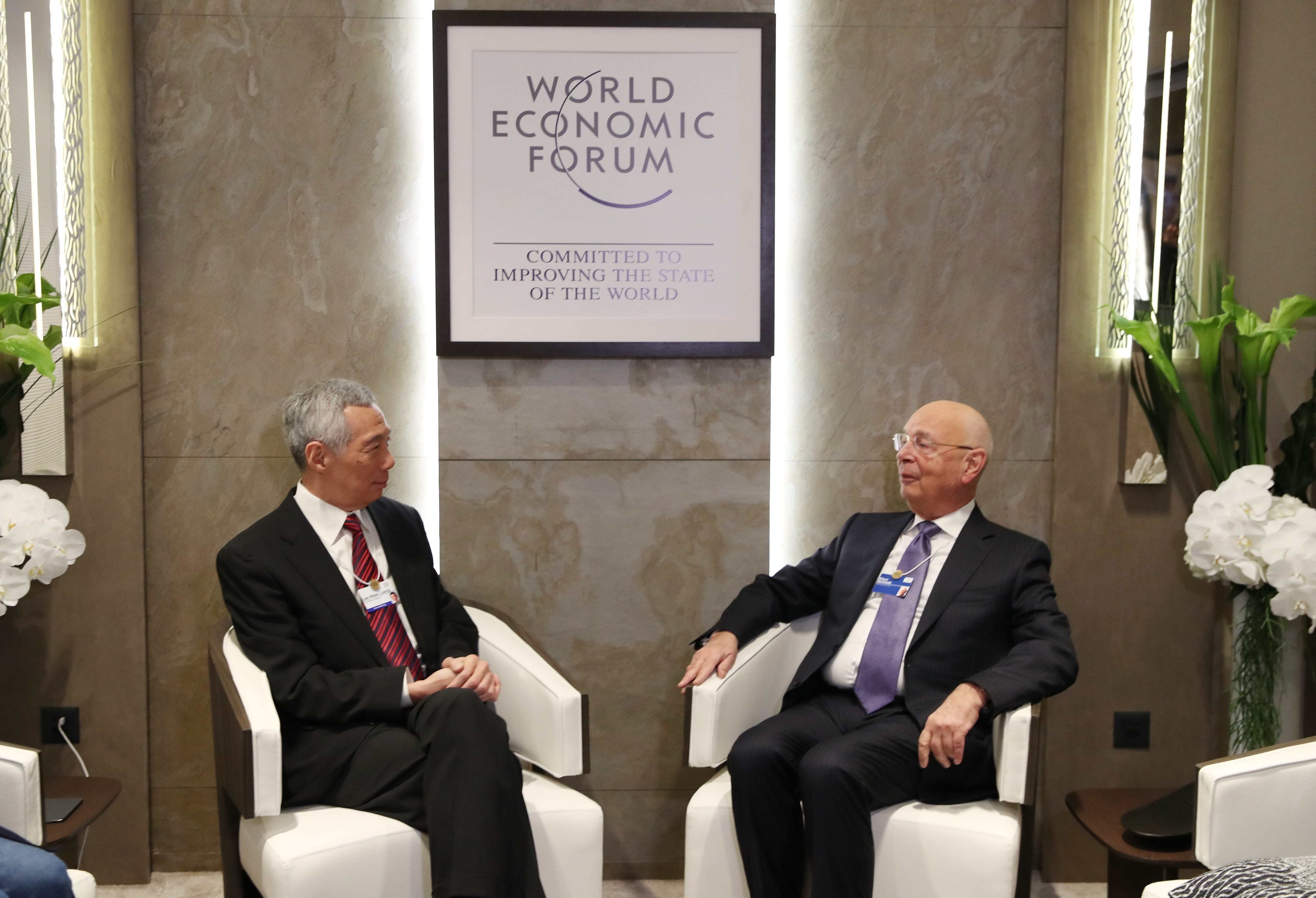 Two men in suits seated in white chairs under World Economic Forum logo.