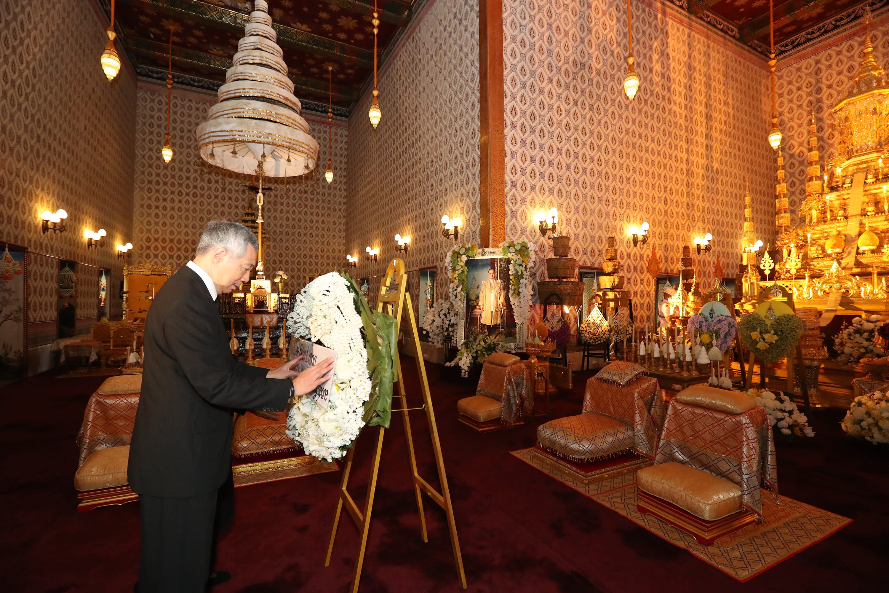 Man places wreath on easel in ornate temple interior with royal portraits and seated cushions.