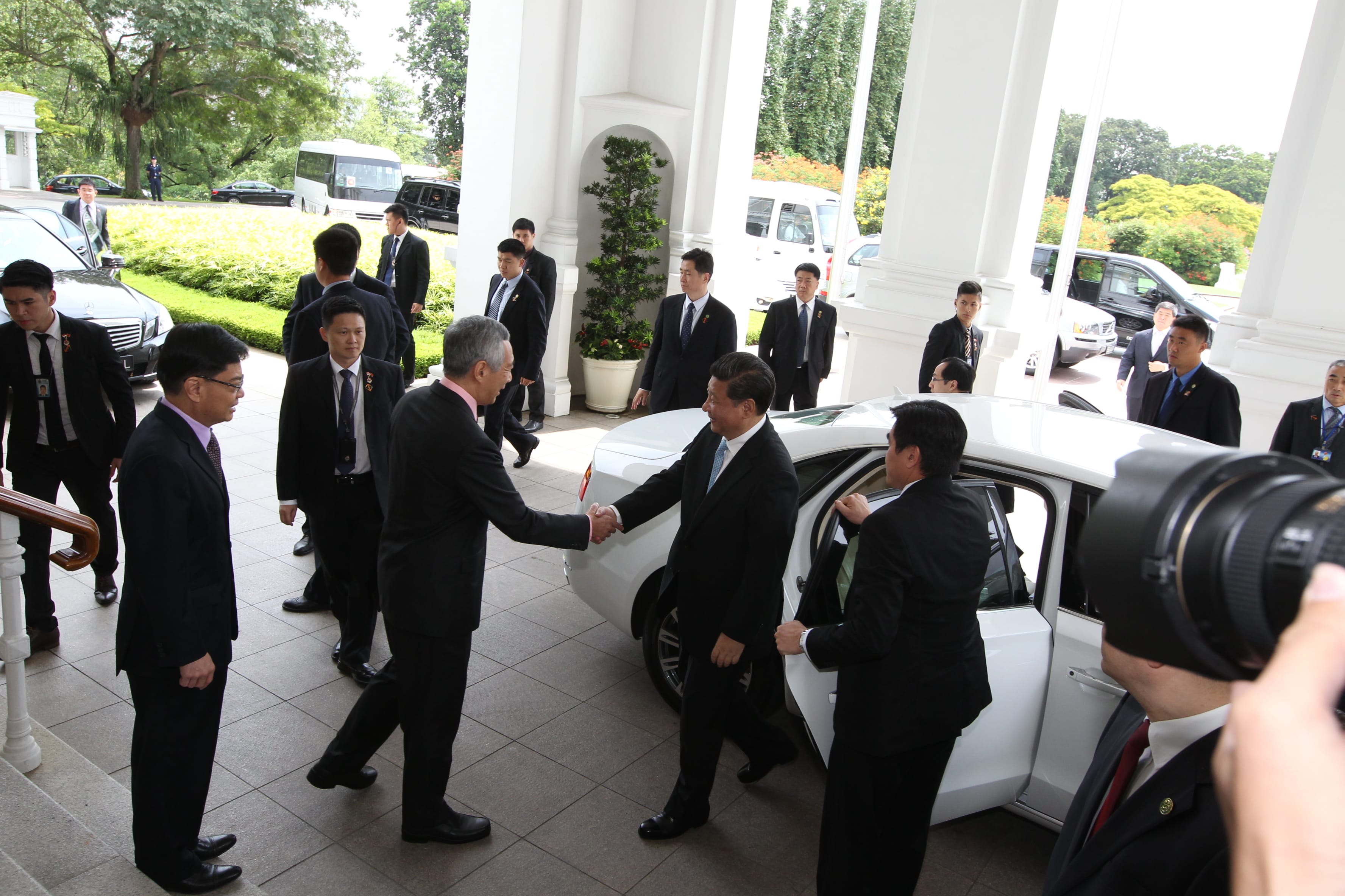Lee Hsien Loong and Xi Jinping shake hands by a white car near a building with white pillars.