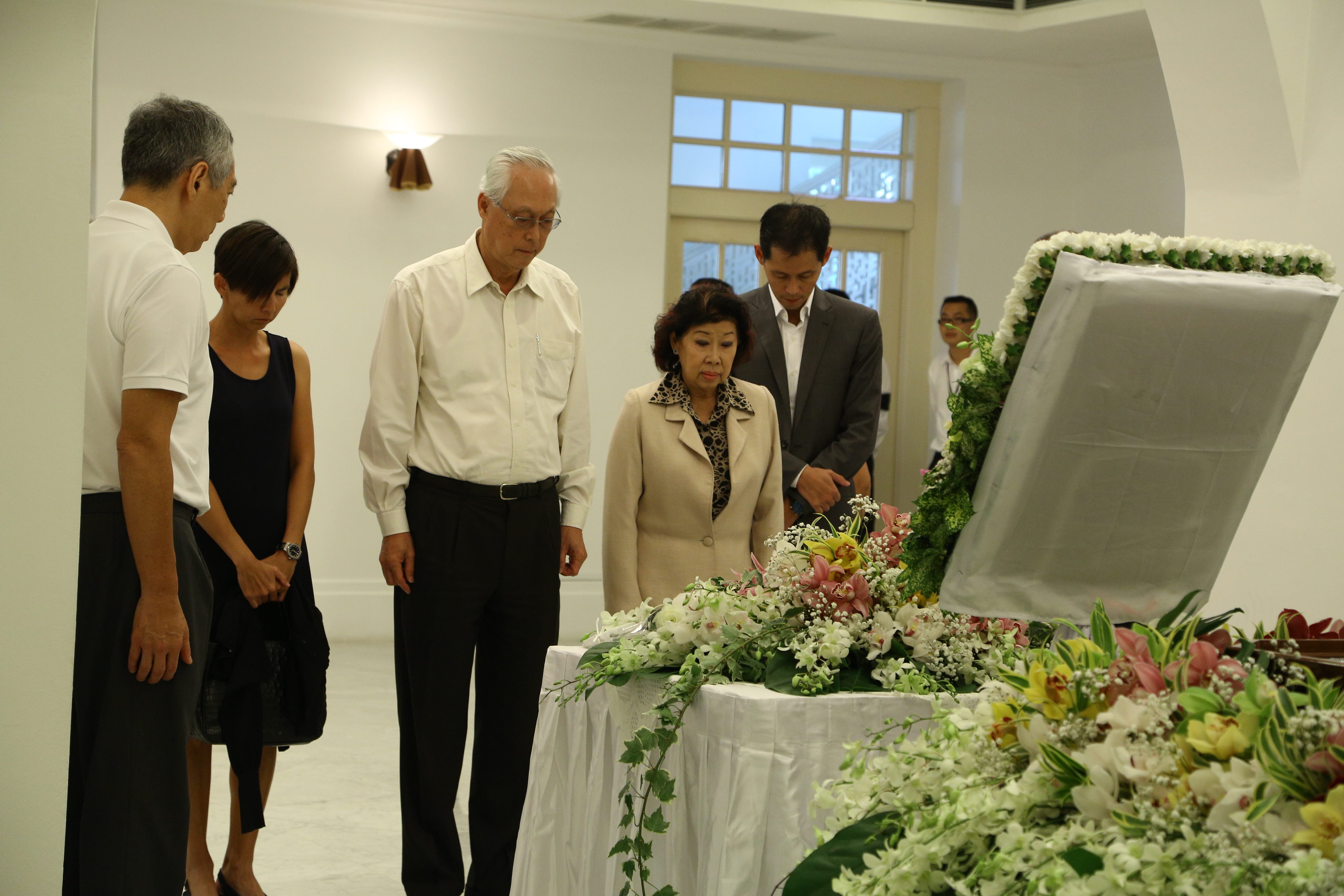 Lee Hsien Loong and others stand at a funeral viewing a flower-laden casket.