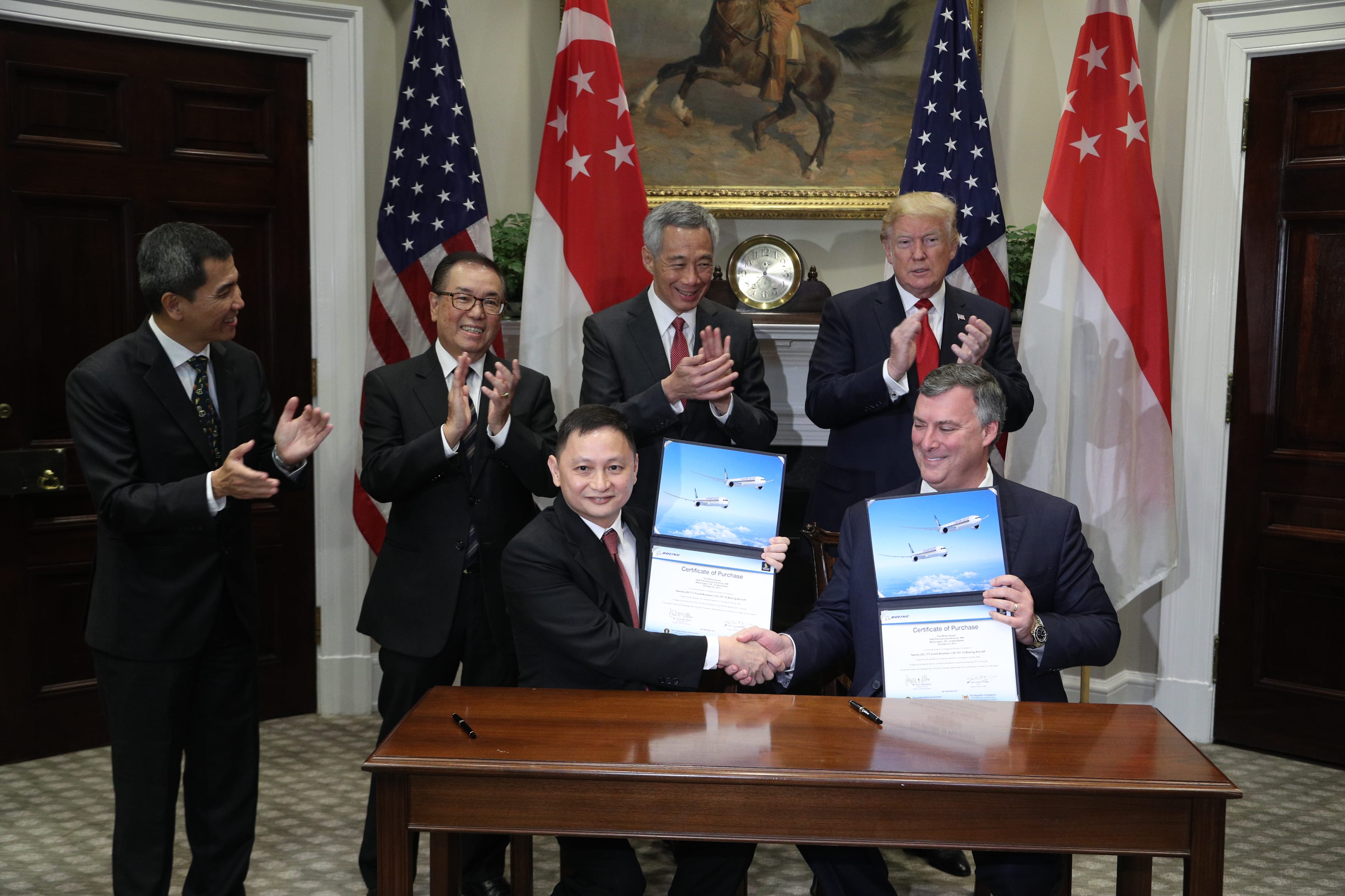 Two men shake hands with Boeing certificates; others, including Trump, applaud before US and Singapore flags.