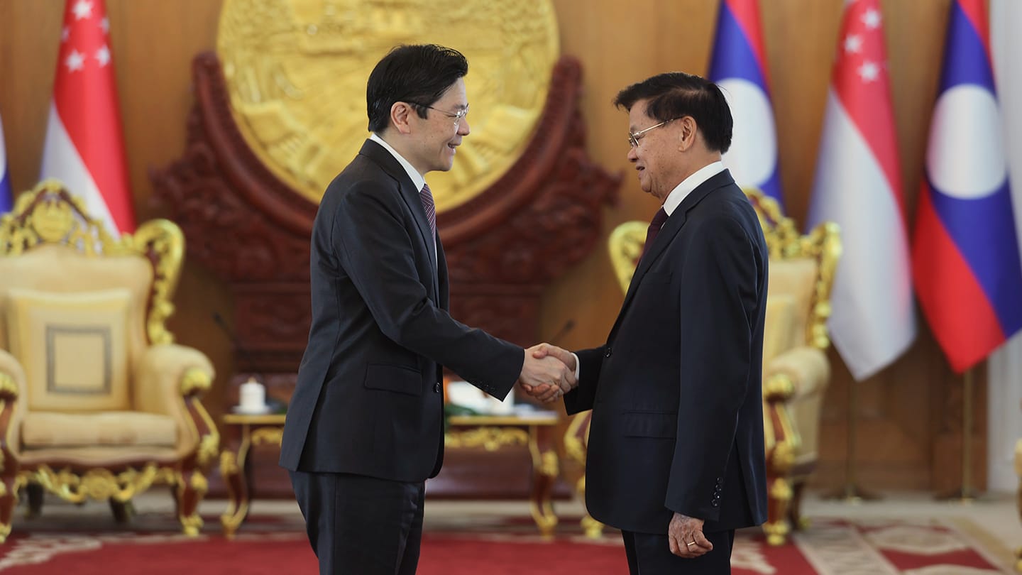 Two men in suits shake hands before flags of Singapore and Laos.