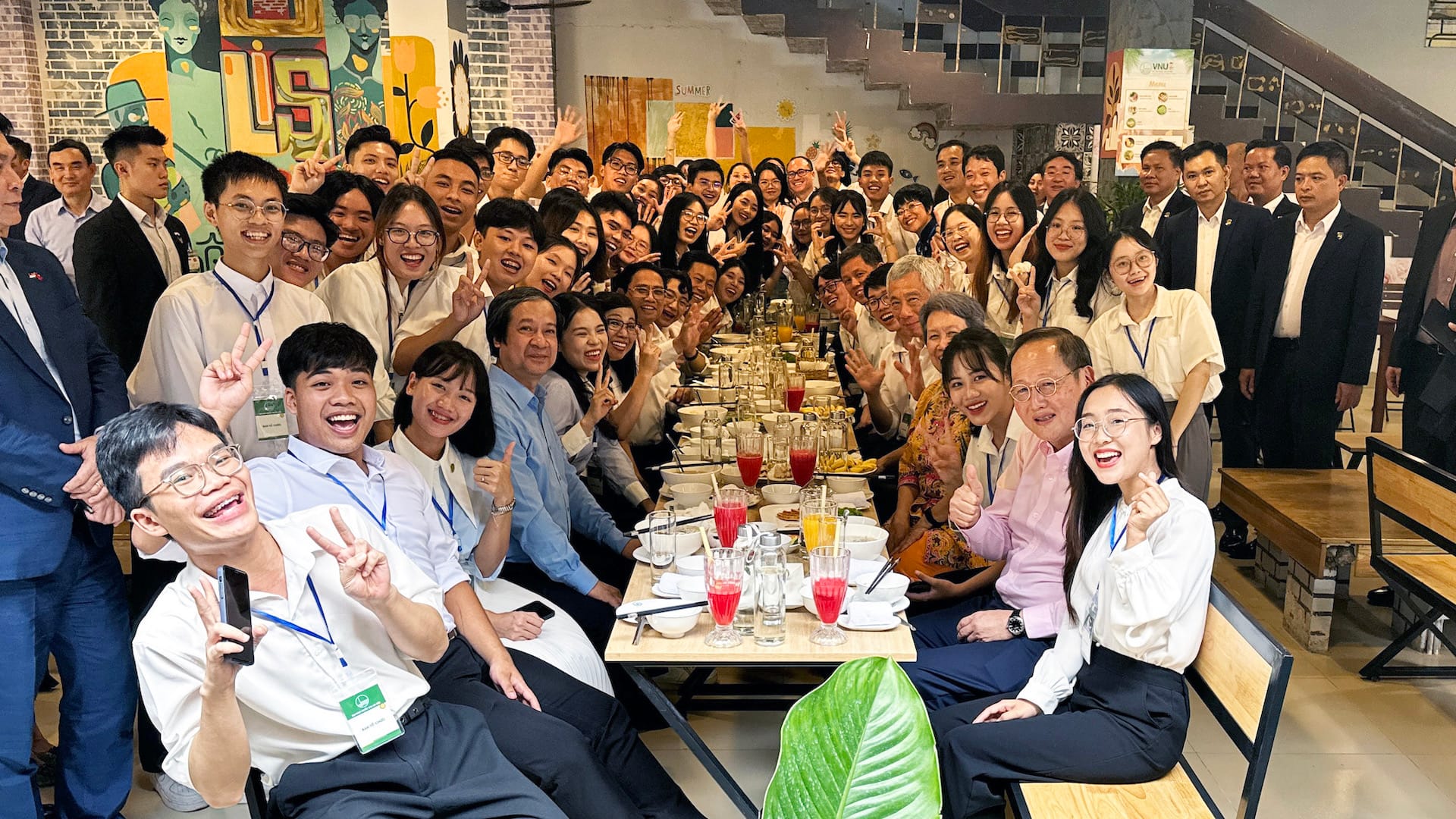 Large group of people gathered around a long table with drinks and plates, waving.