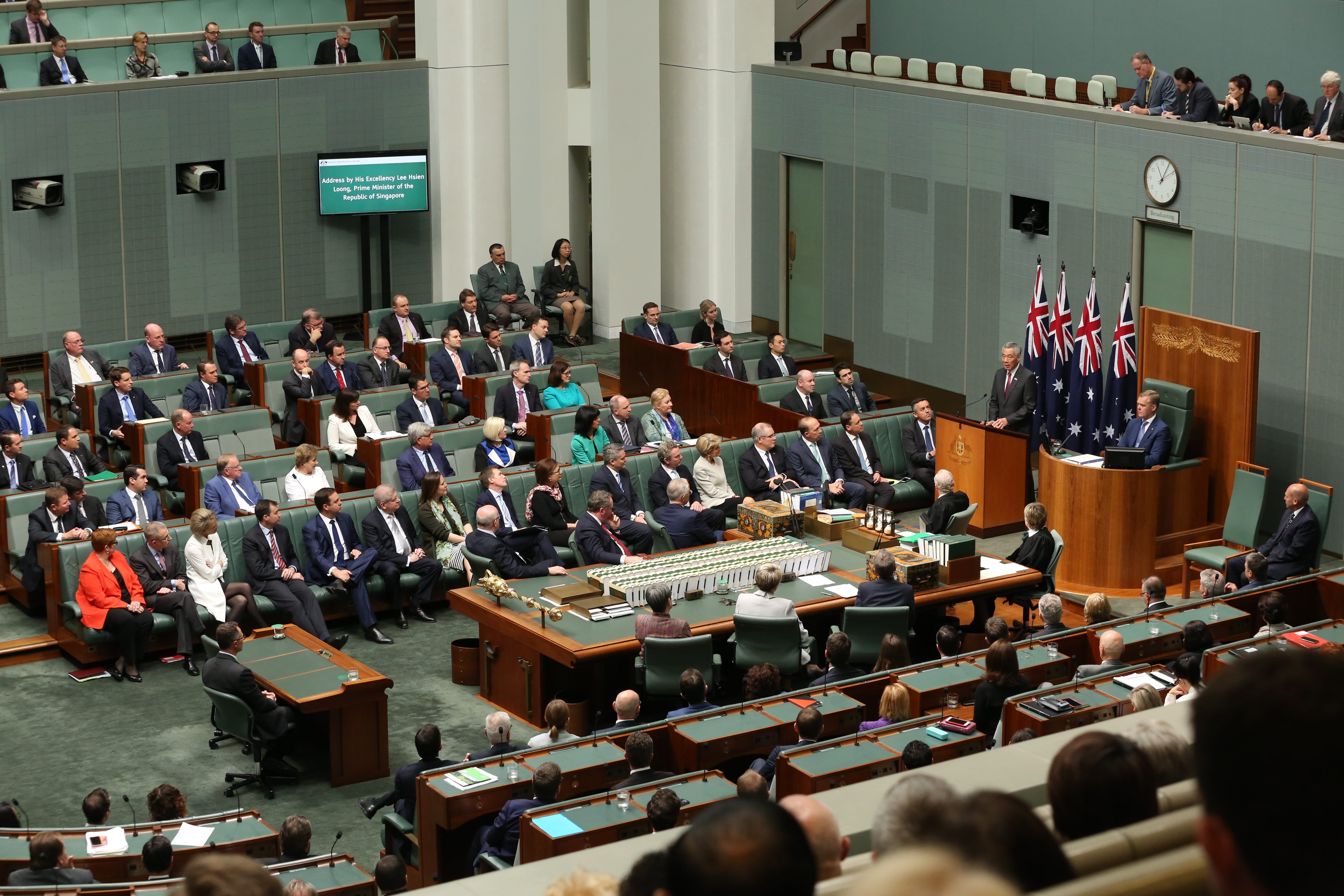 Large chamber with people seated, man at podium with Australian flags.