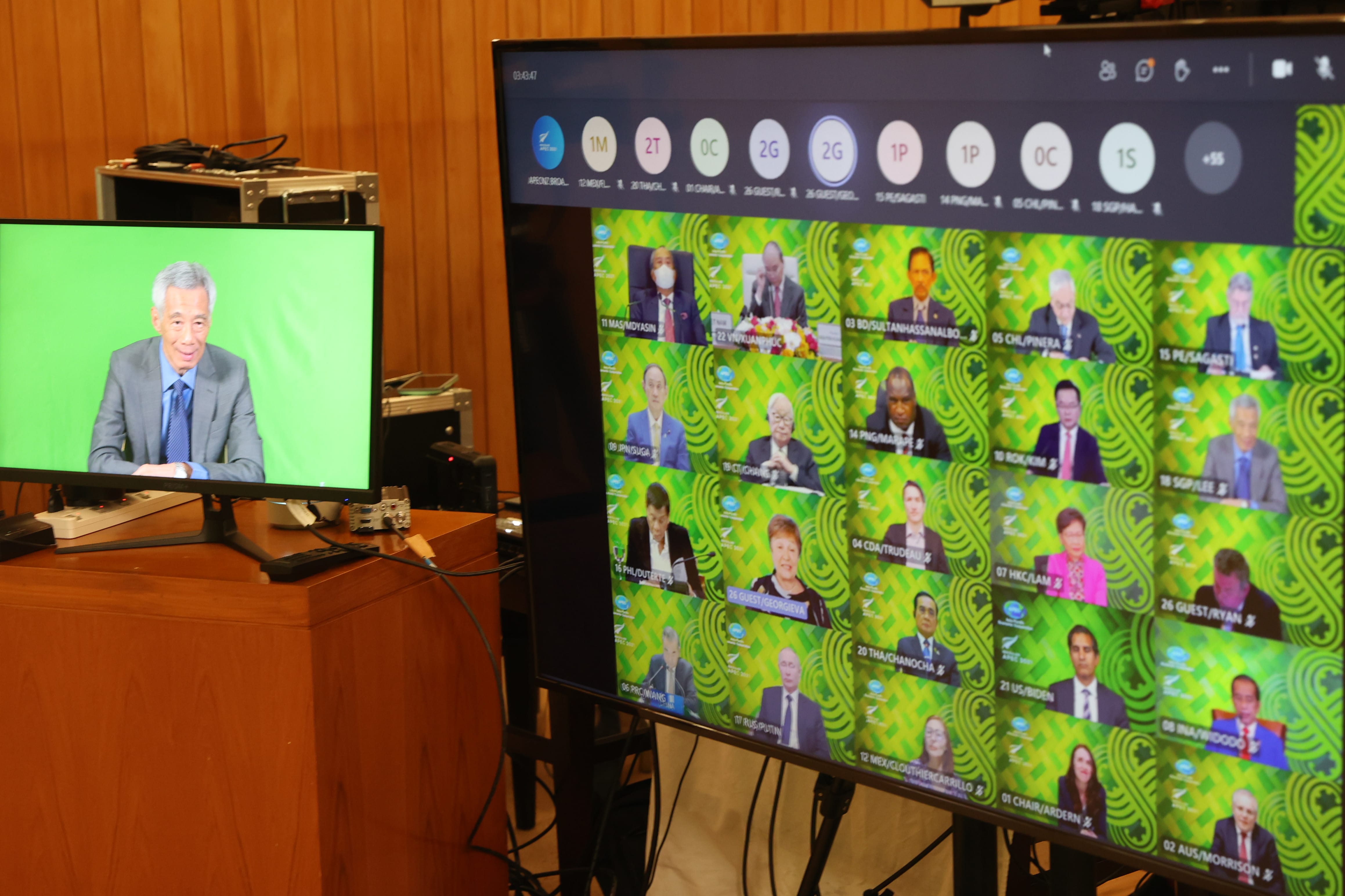 Two monitors: one with Lee Hsien Loong on a green screen, the other showing a virtual APEC meeting.