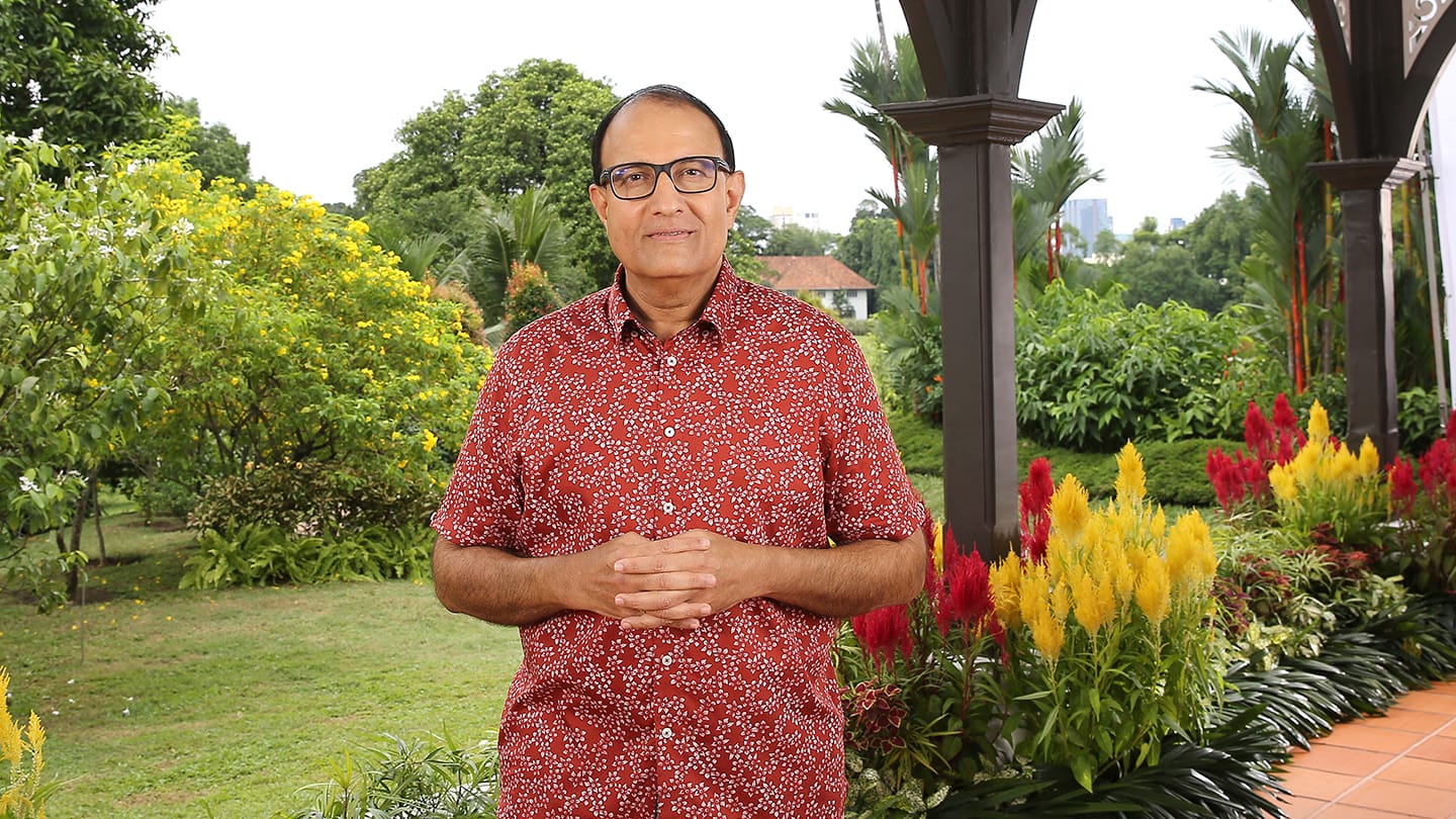 Man in glasses, red floral shirt, hands clasped, stands in a lush garden.