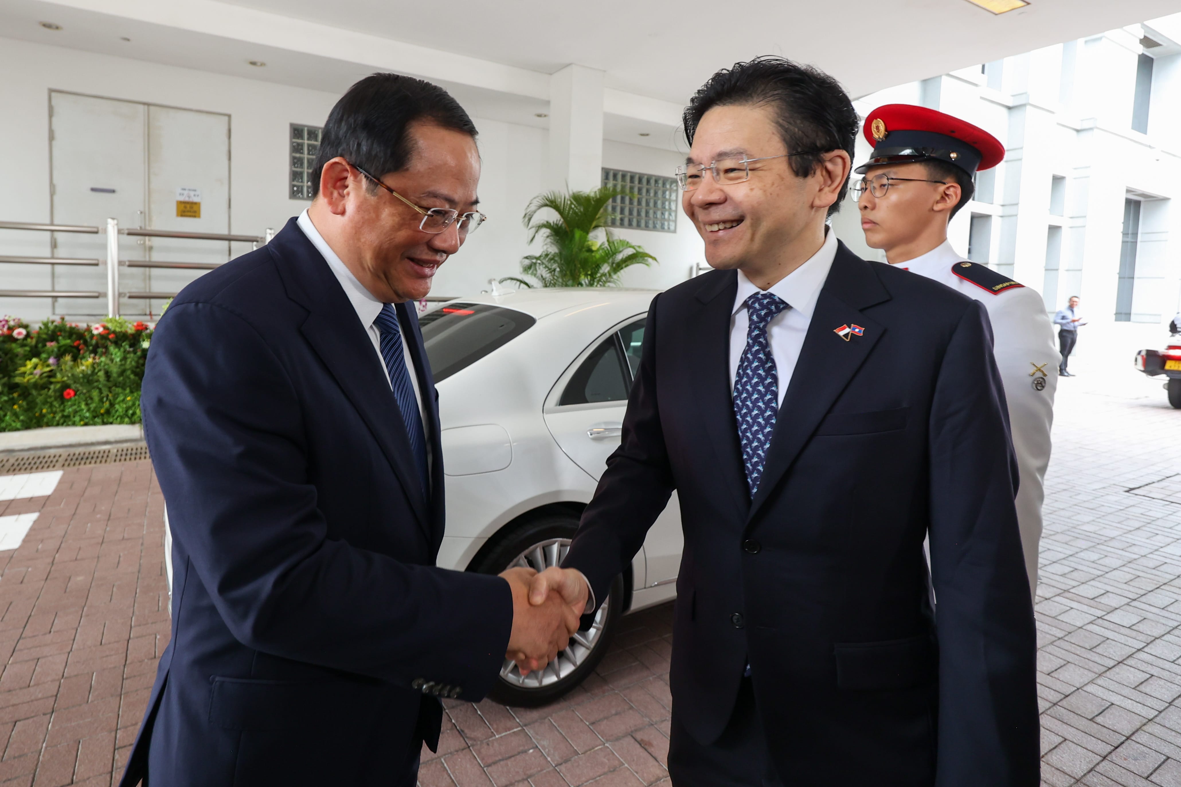 Two men in suits shaking hands; a white car in the background; another man in uniform stands by.