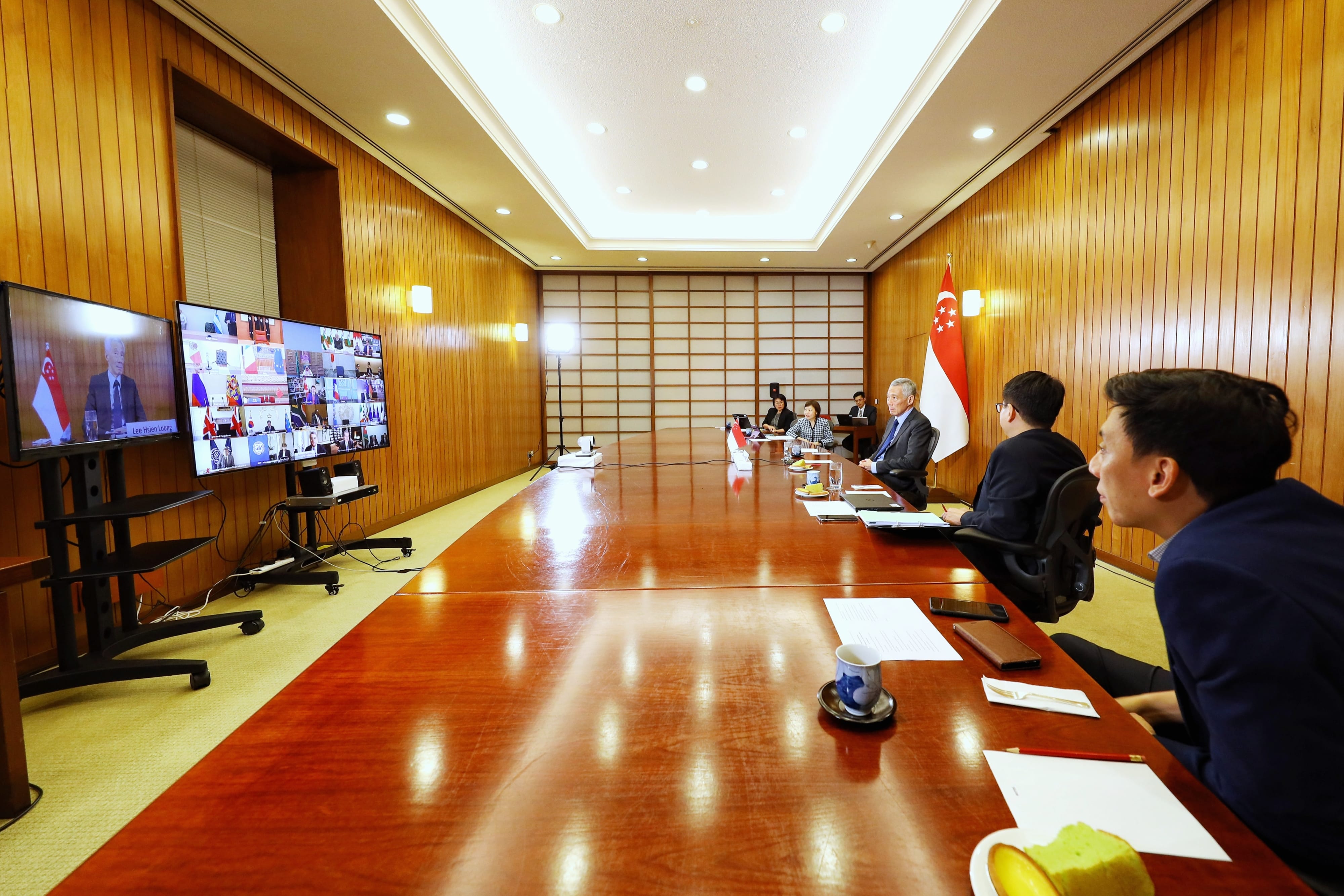 Conference room with Lee Hsien Loong on screen, Singapore flag, and people at table during video call.