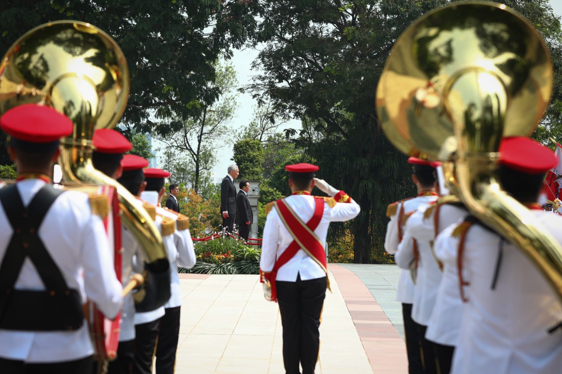 Military band in red hats and white uniforms, one saluting, with two men in suits in the background.