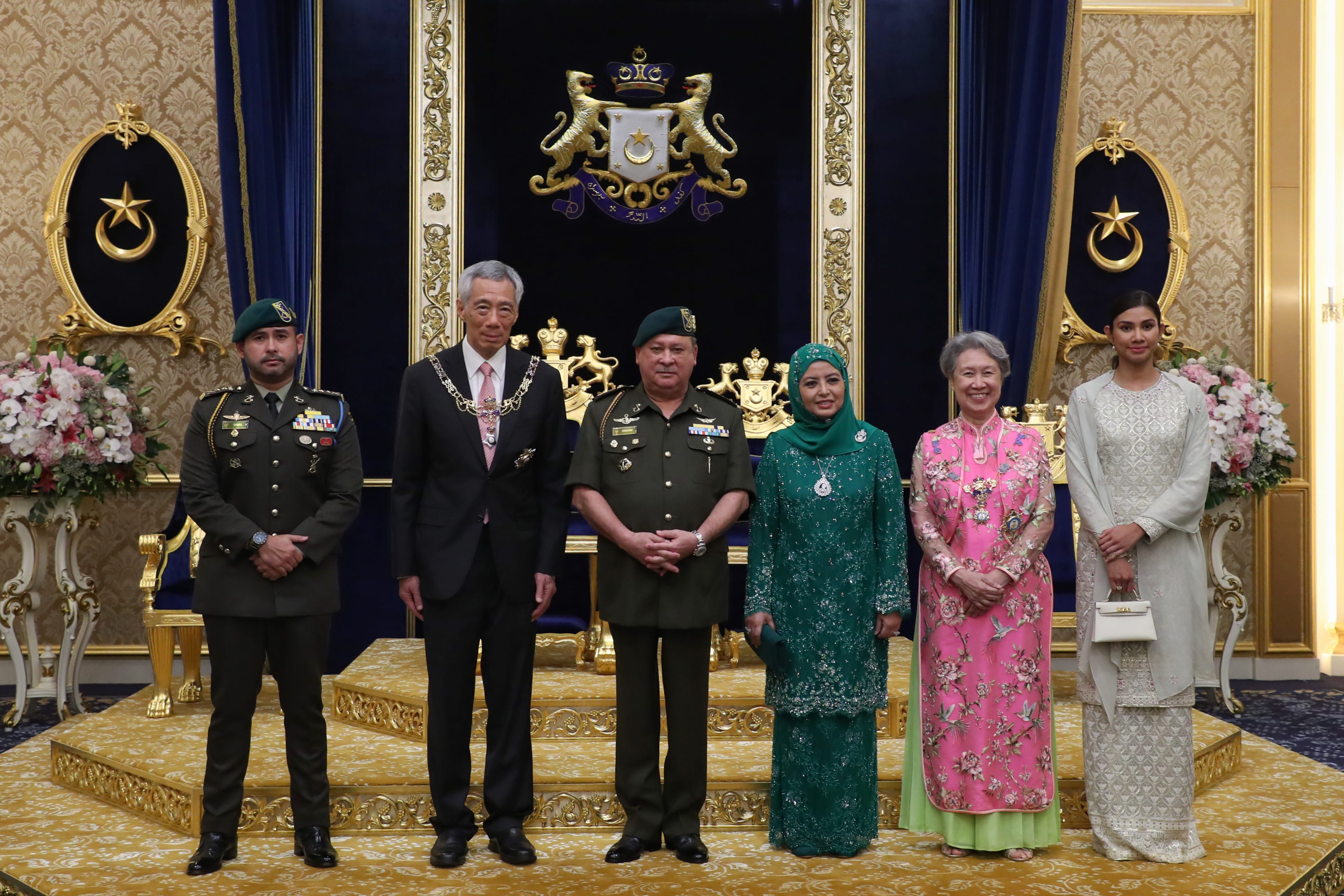 Six people in formal attire standing on a gold patterned platform before royal emblem.