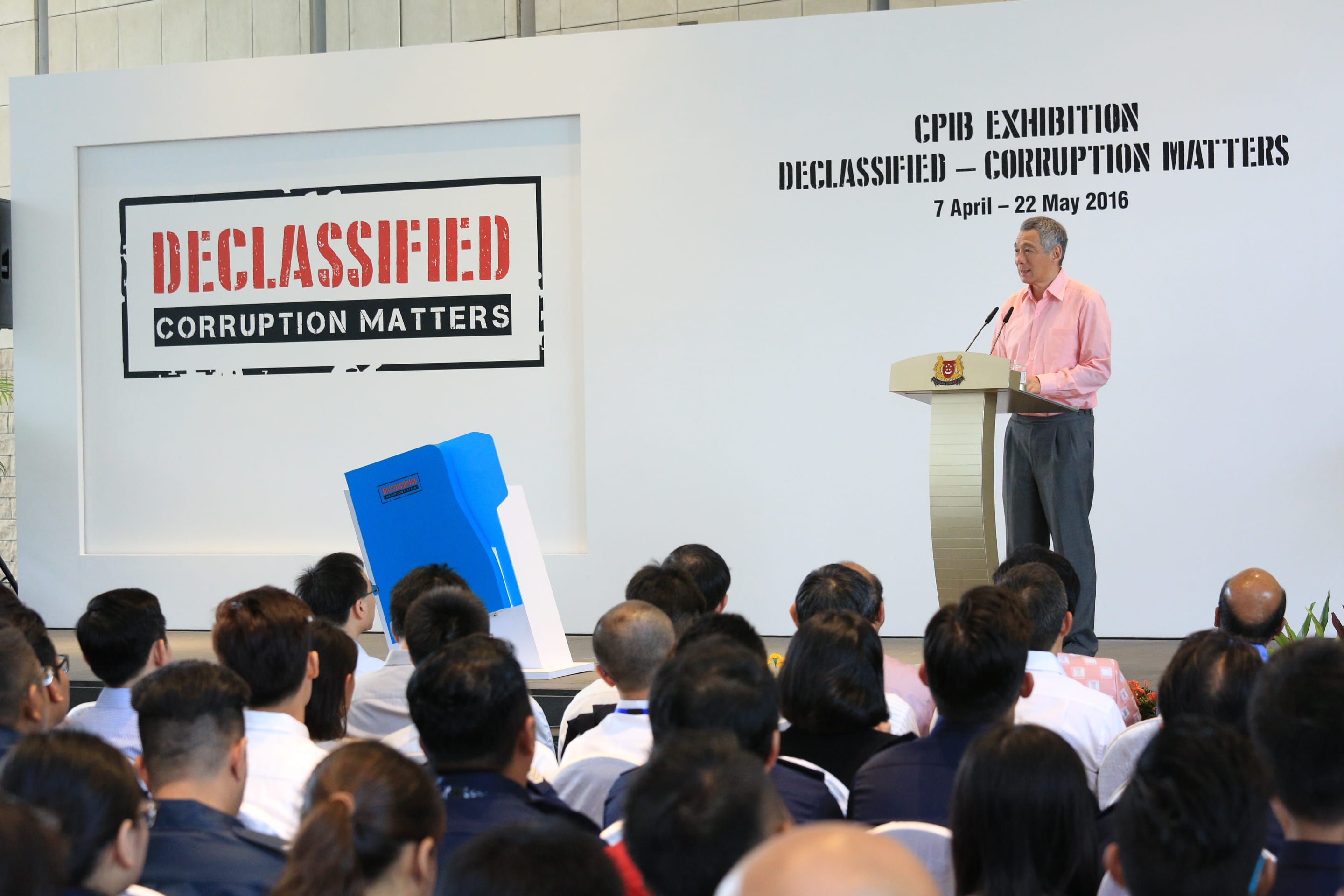 Lee Hsien Loong at podium giving a speech in front of CPIB "Declassified" exhibit.