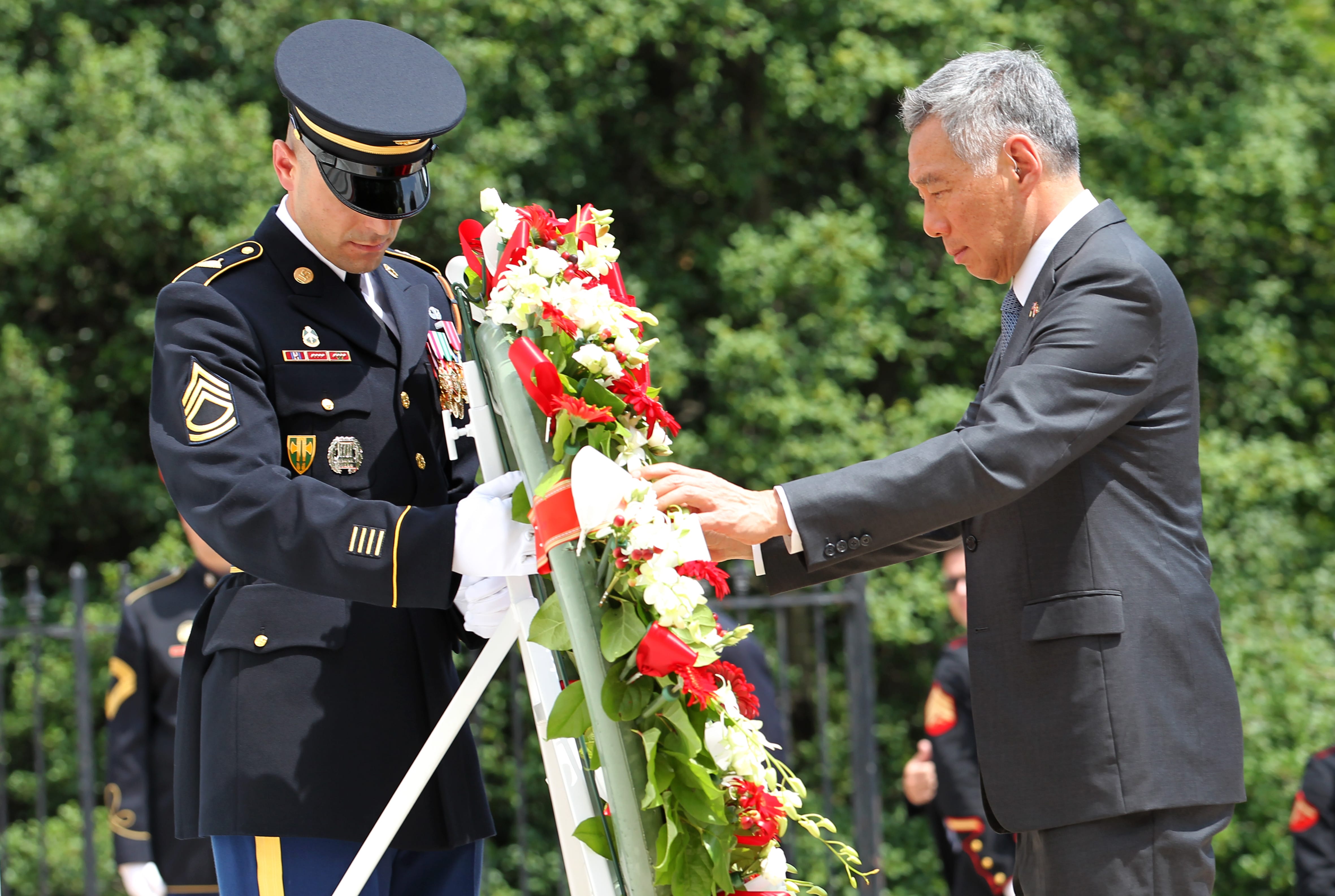 Soldier and man in suit laying red and white flower wreath against greenery.