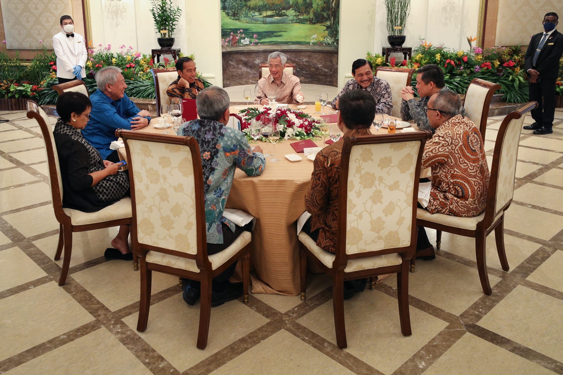 Group of people at round table with floral centerpiece, wait staff nearby.