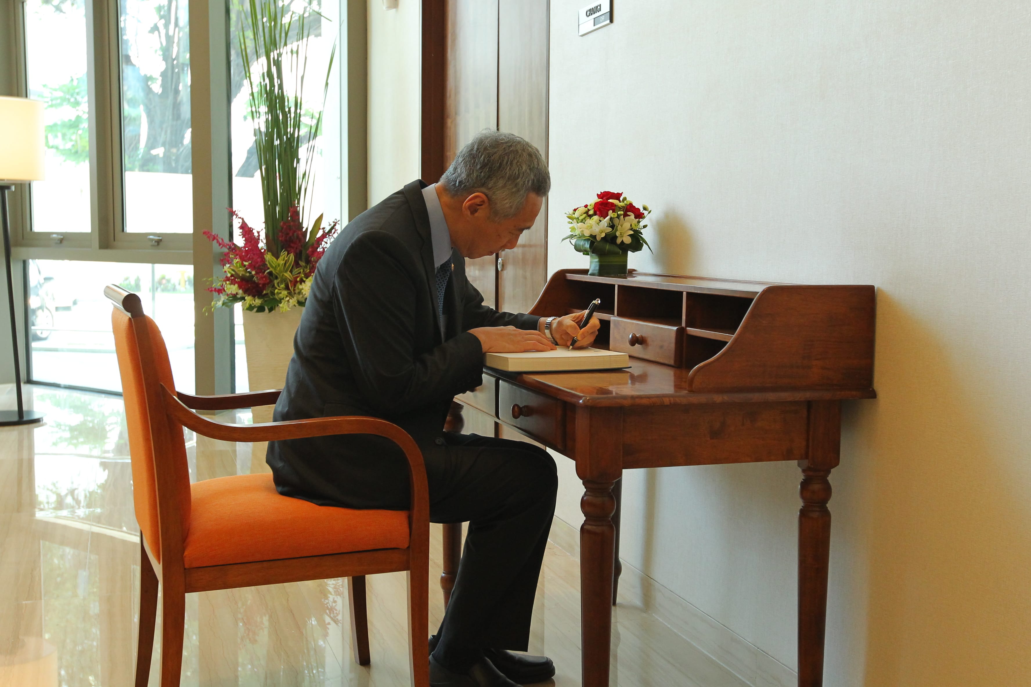 Lee Hsien Loong signing a book at a wooden desk, seated in an orange chair.