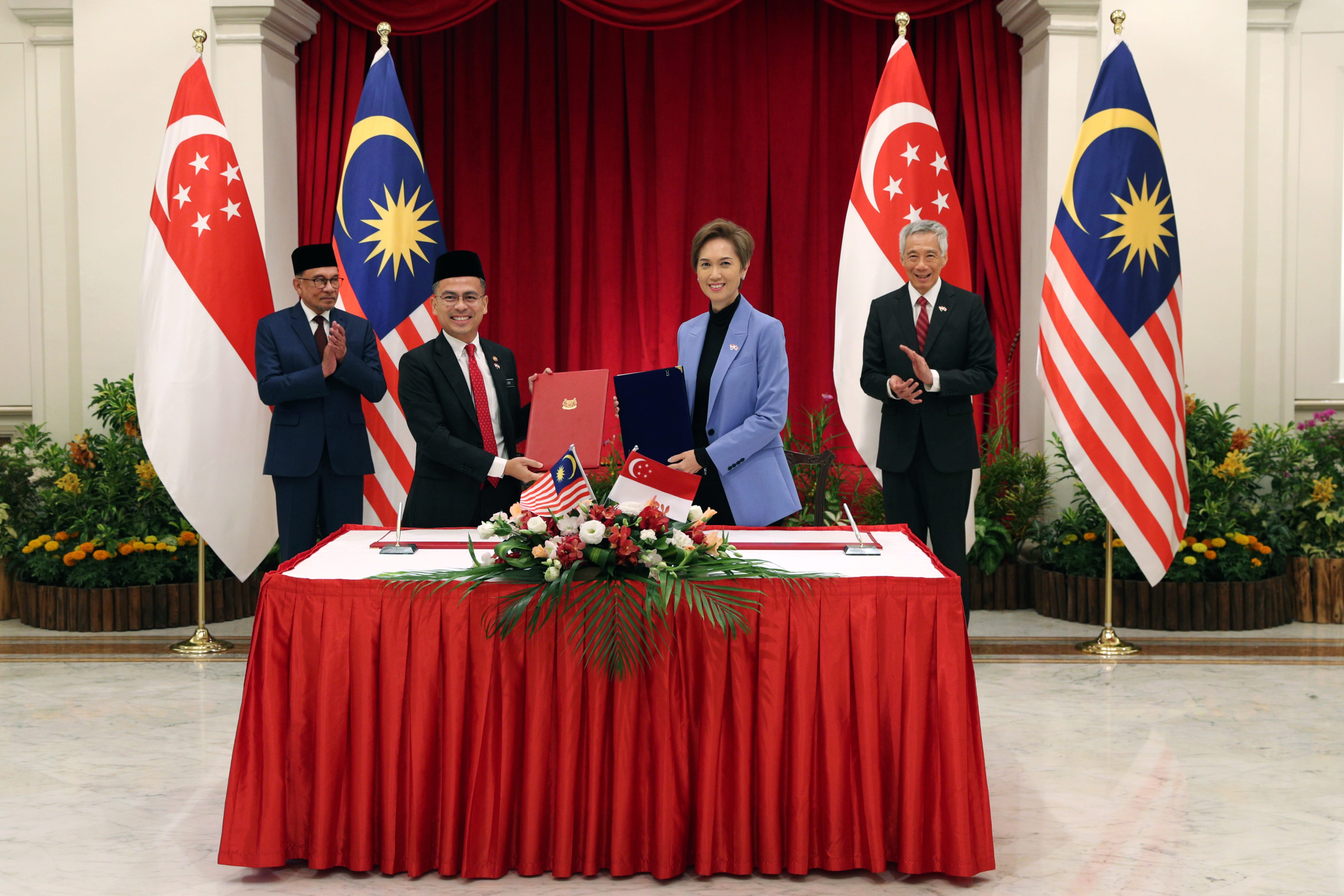 Two leaders signing papers on a red-draped table with Malaysia and Singapore flags. Men clap.
