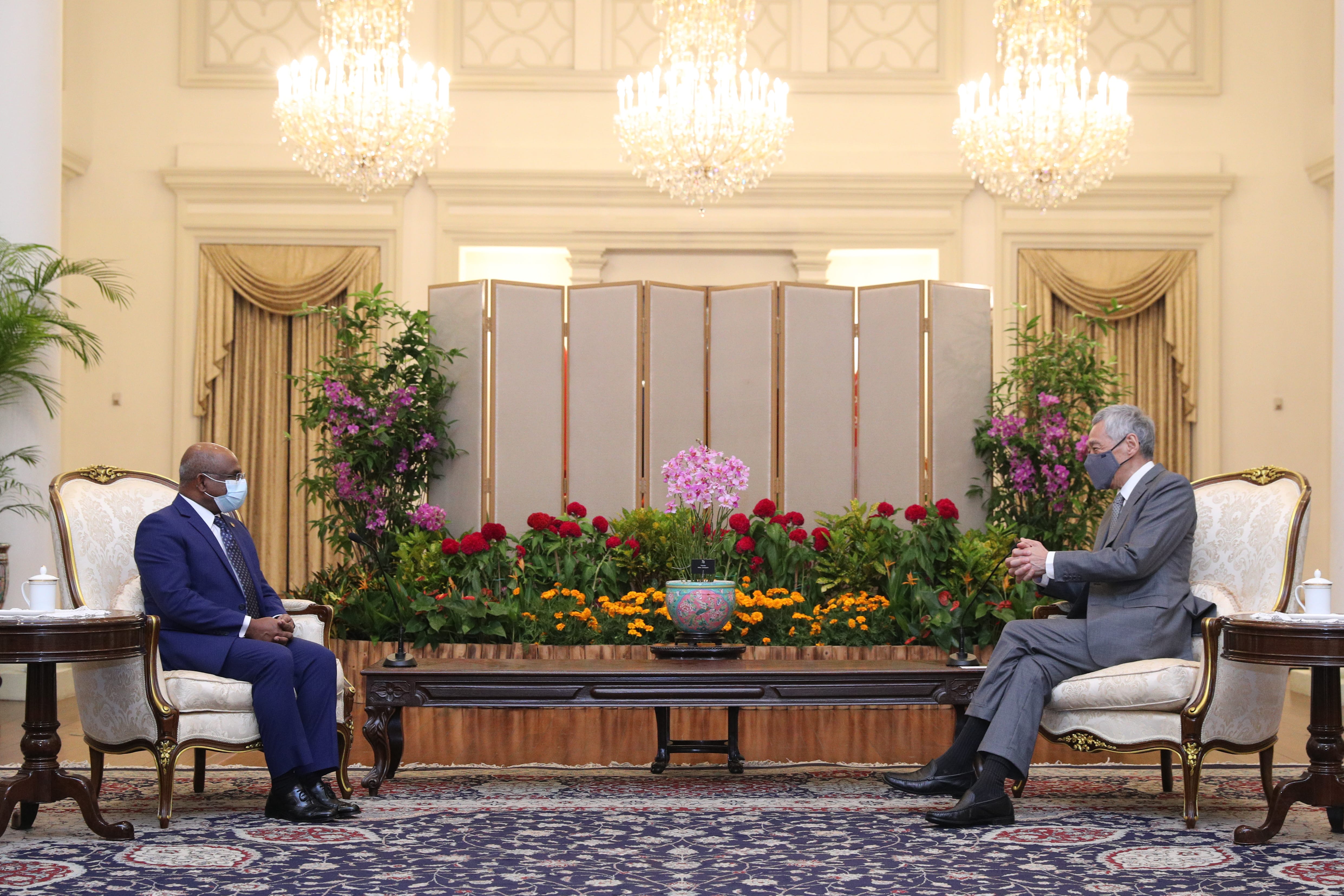 Two people seated in ornate chairs, separated by flowers and a screen. Both are wearing masks.