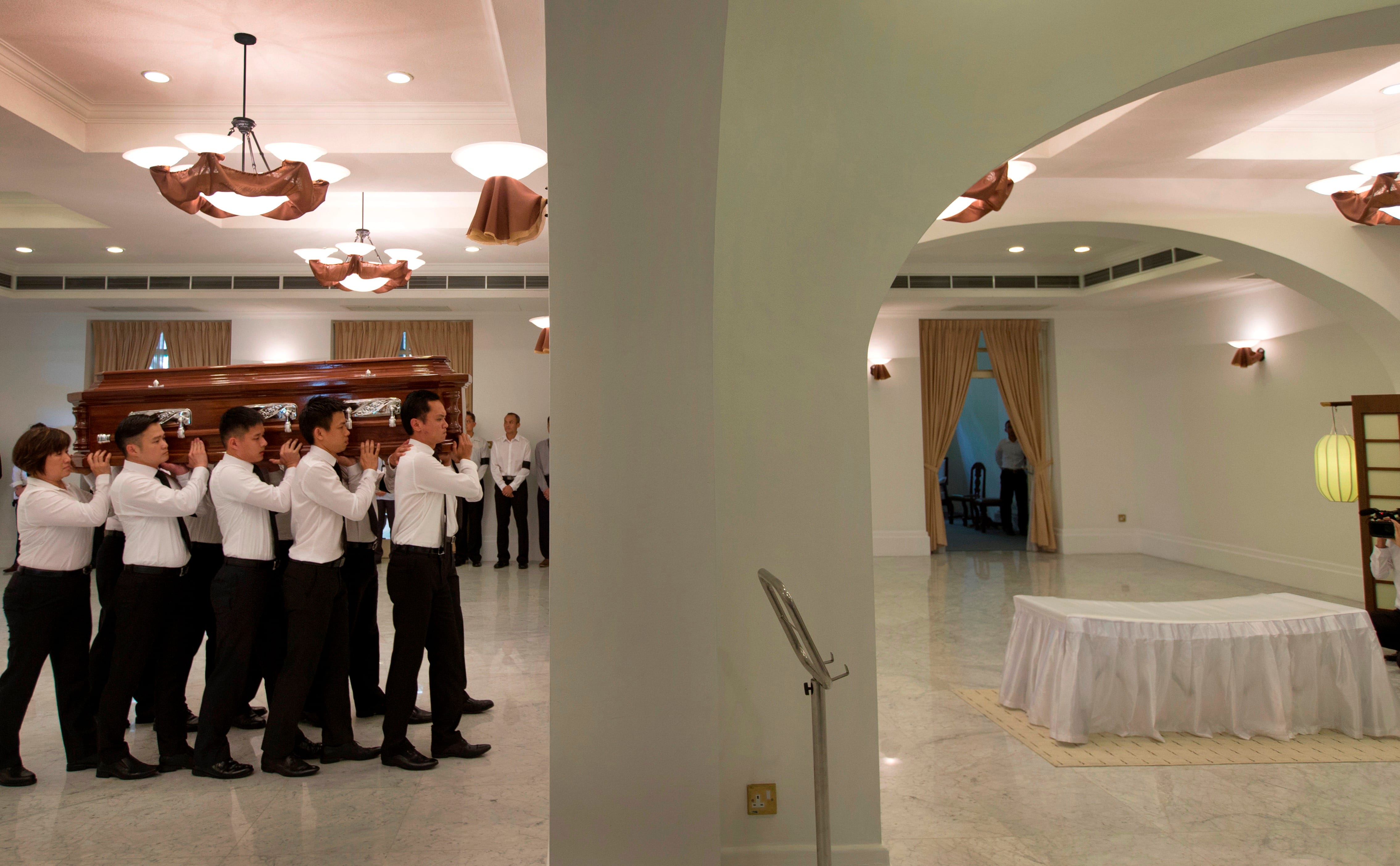 Pallbearers carry a wooden casket inside a building. A covered table is nearby.