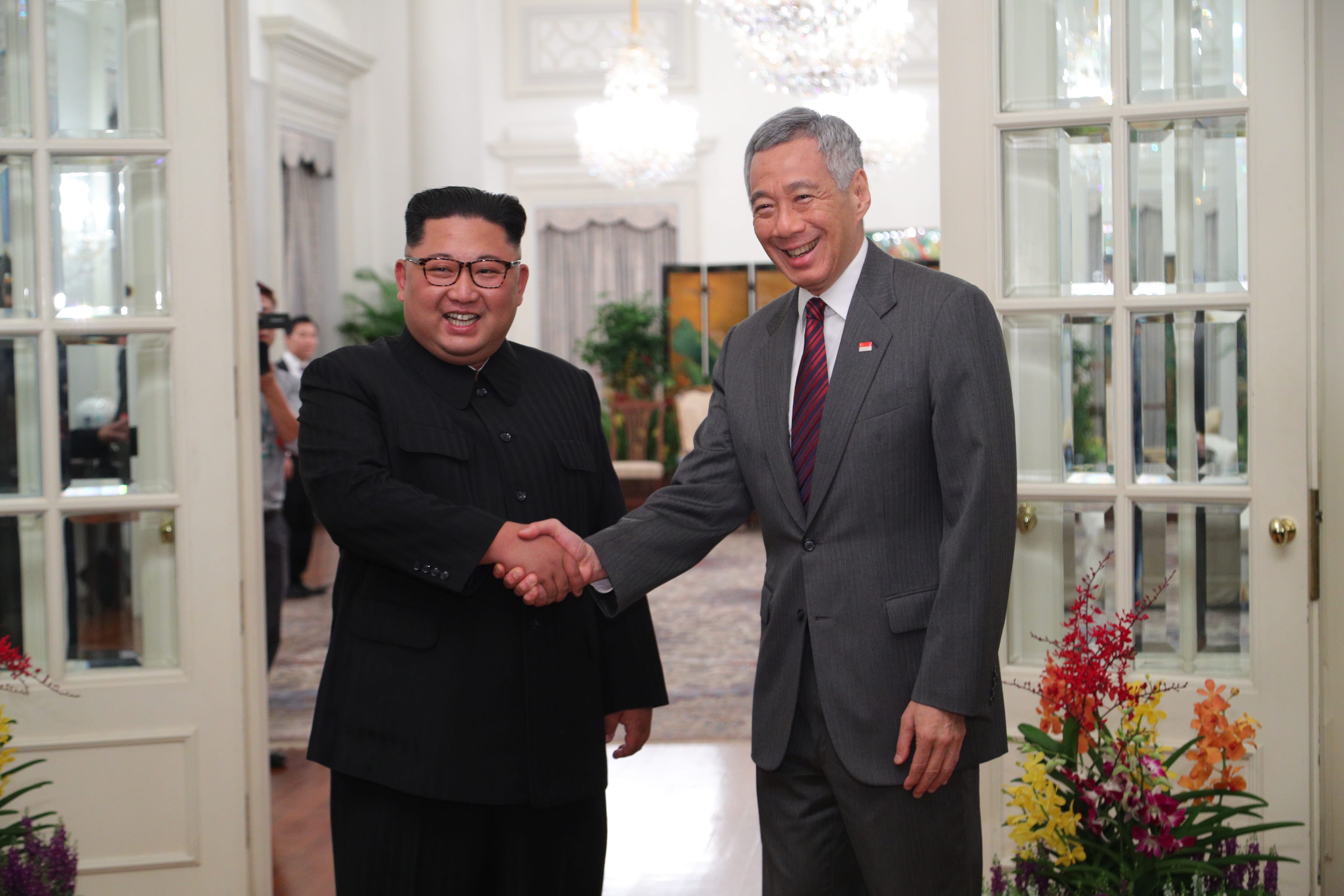 Kim Jong-un and Lee Hsien Loong shake hands between white glass doors.