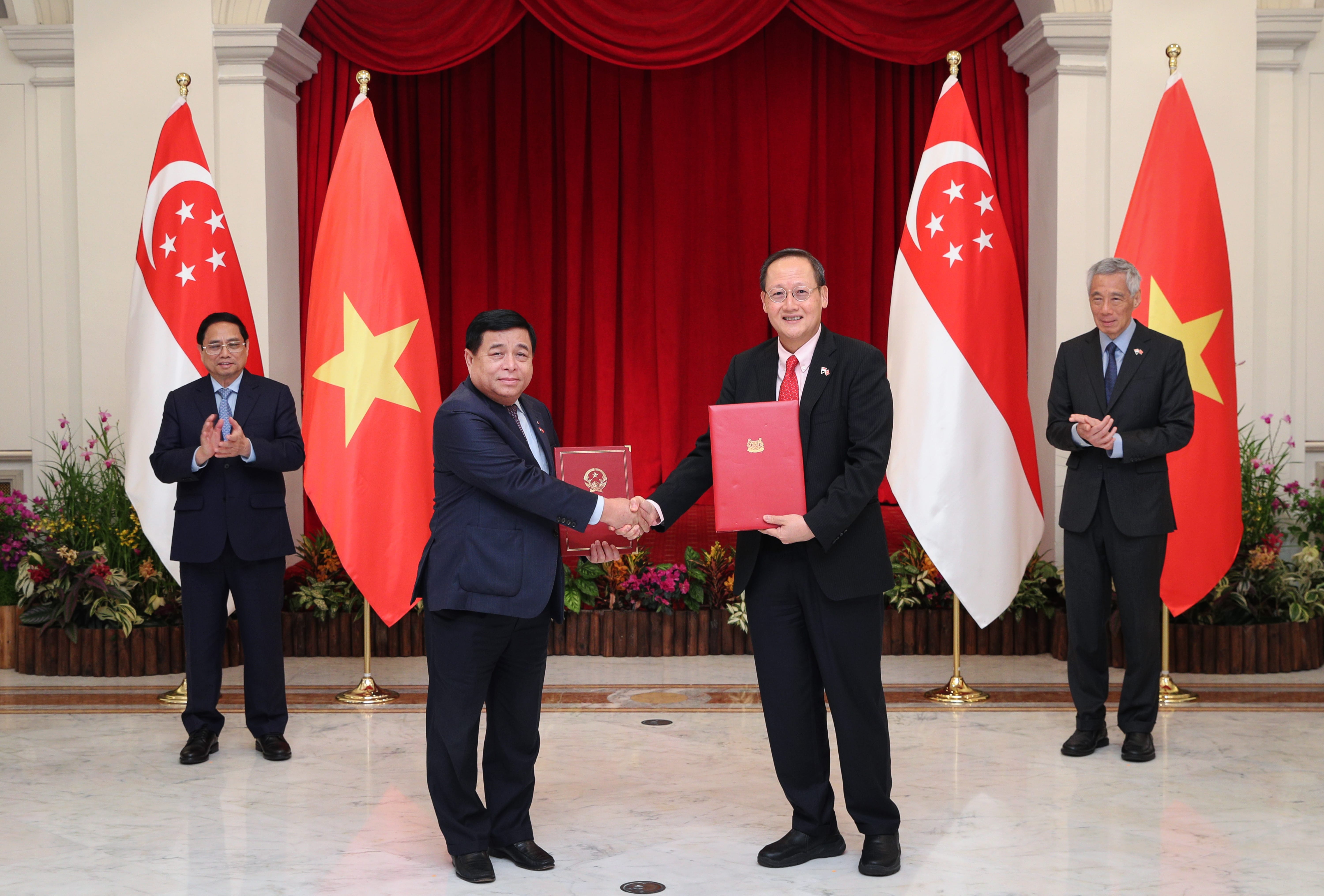 Two men shaking hands, each holding a red folder, flanked by Singapore and Vietnam flags.