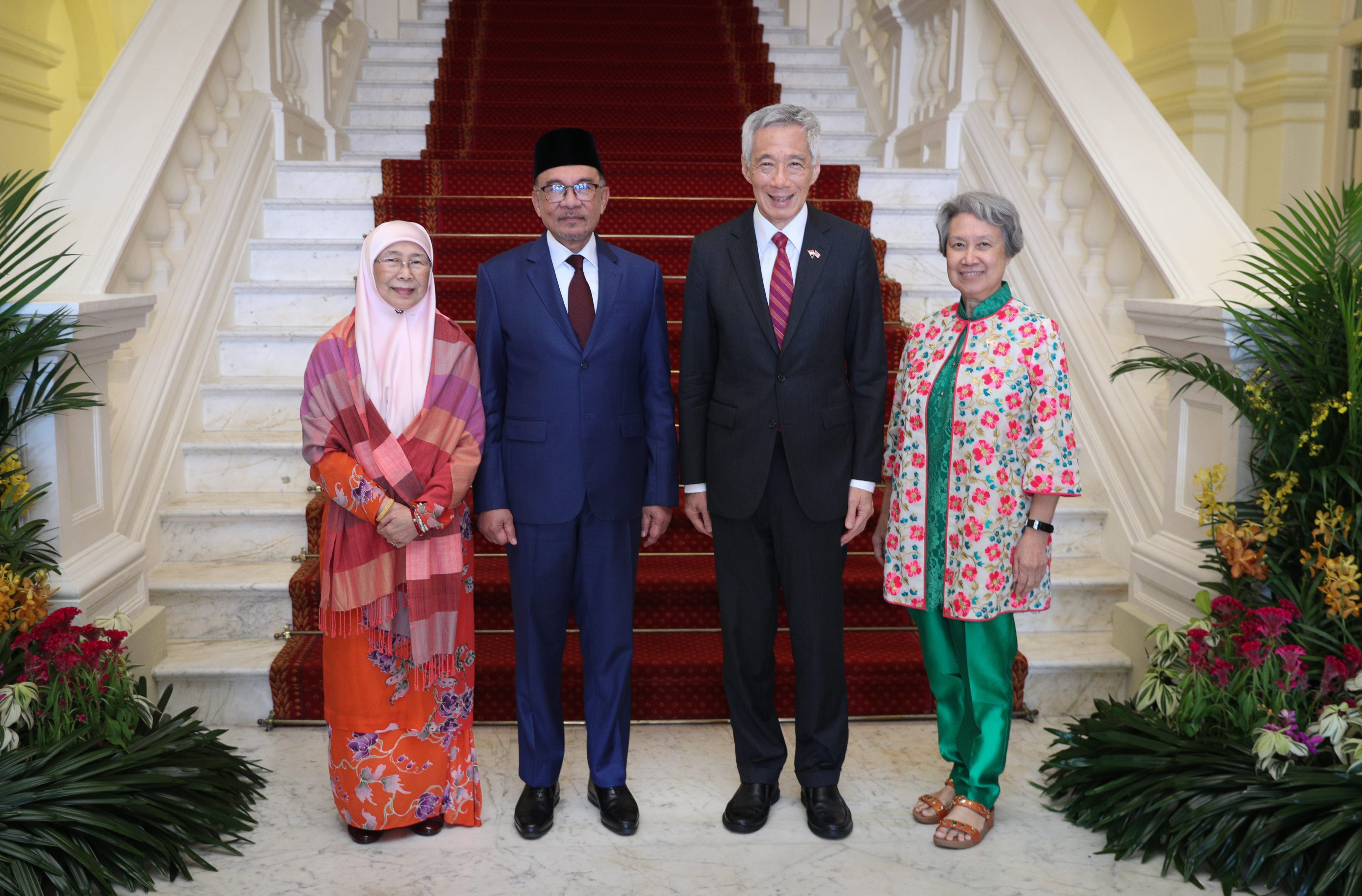 Four people stand on a marble floor before stairs with a red runner. They are formally dressed.