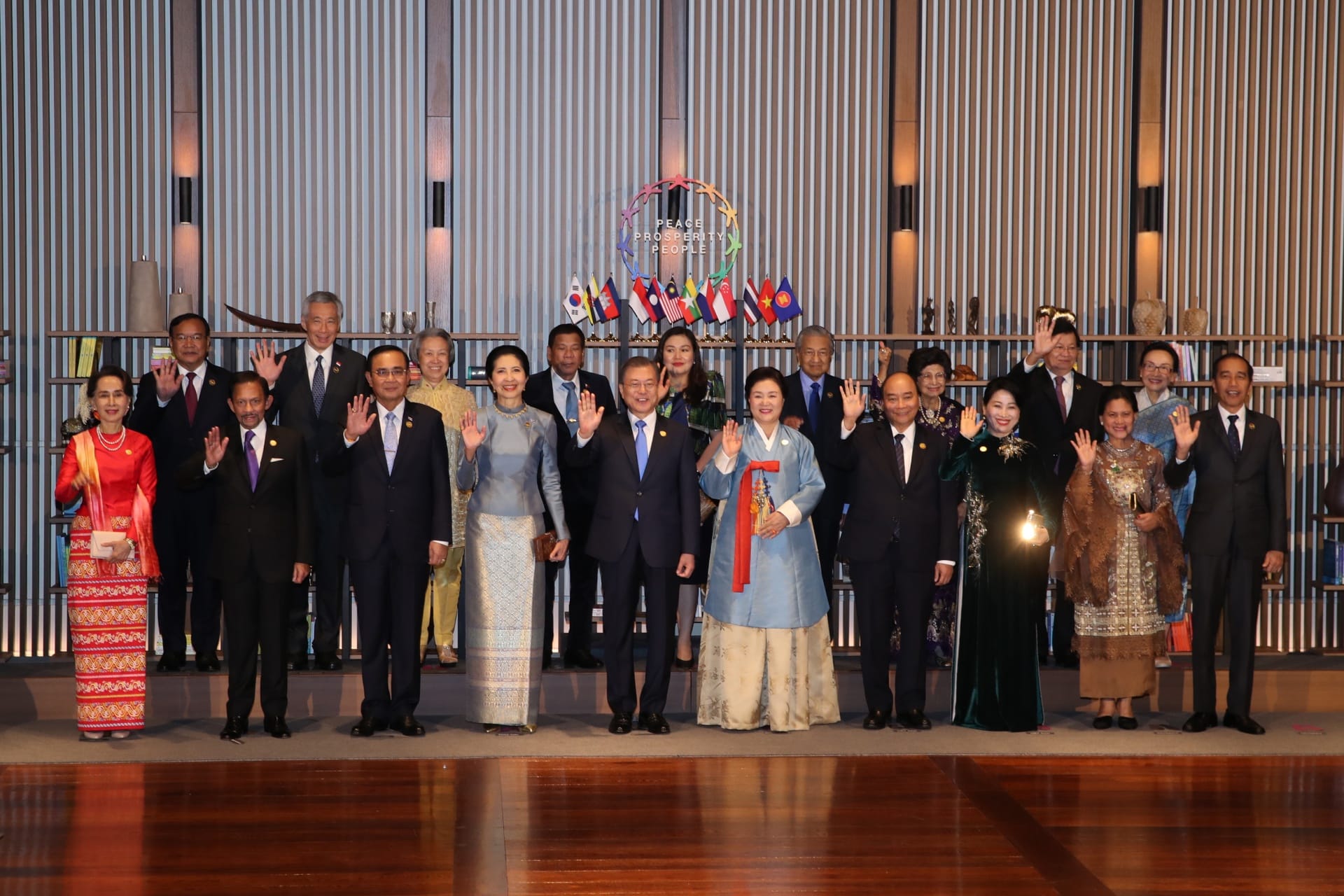 Large group of dignitaries waving in formal attire, flags and logo "Peace Prosperity People" in background.