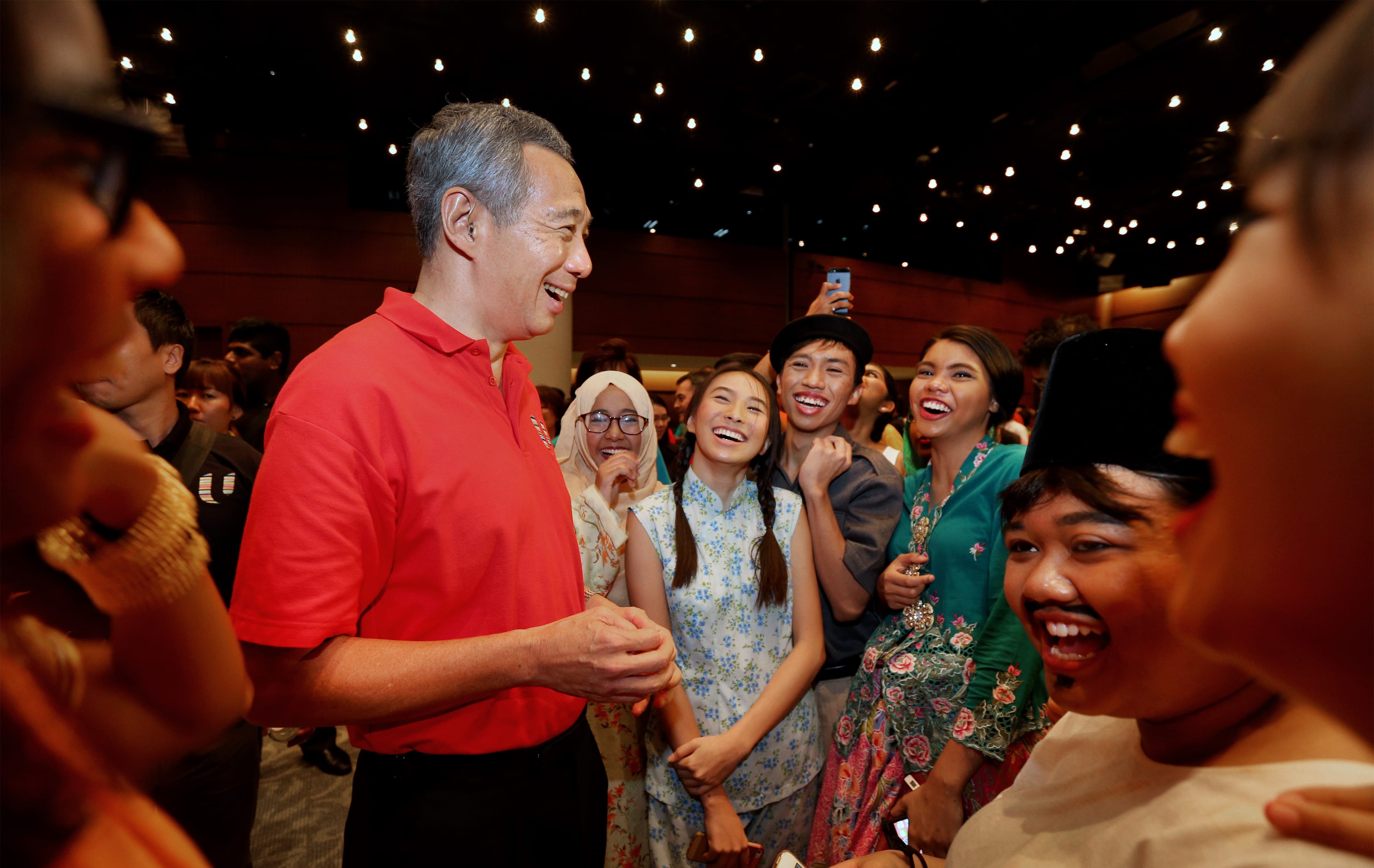 Lee Hsien Loong in red polo shirt, smiling with a laughing crowd, some in traditional clothing.