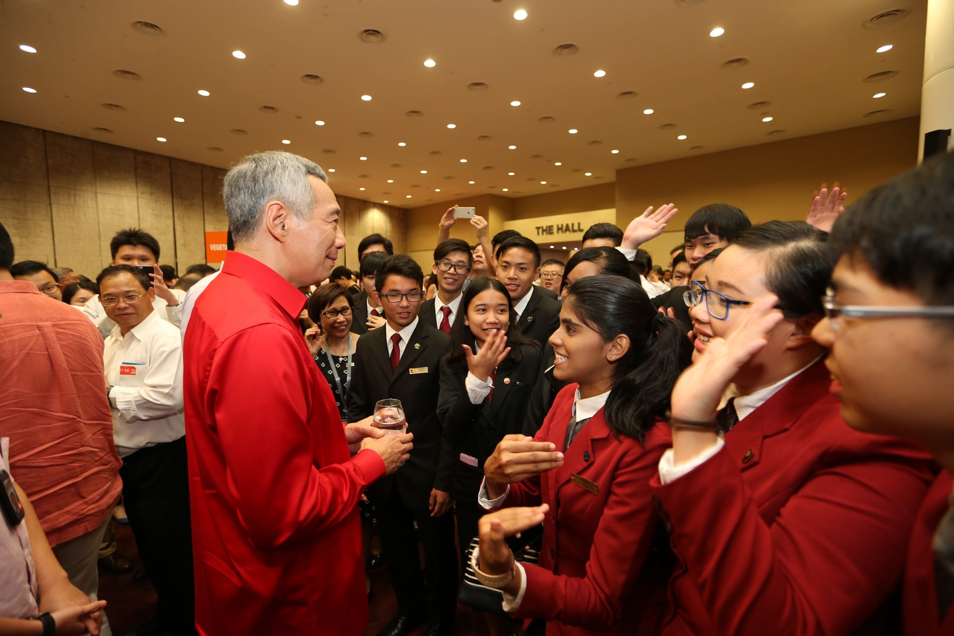 Singapore's PM Lee Hsien Loong surrounded by students in red and black uniforms, holding glass.