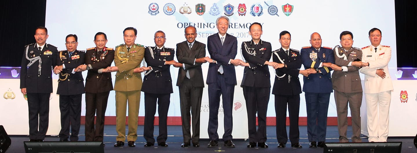 People in uniforms and business suits stand hand-in-hand on a stage with national emblems in the background.