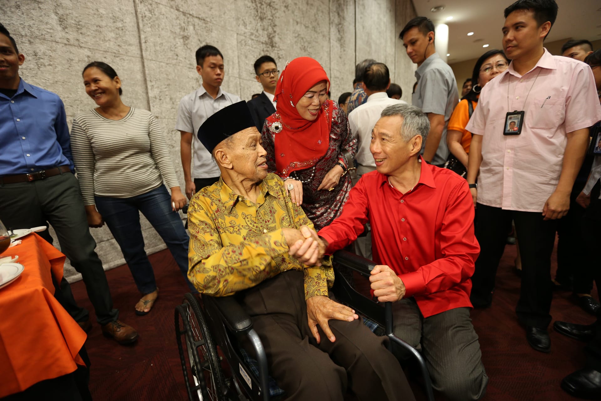 Older man in wheelchair shakes hands with Lee Hsien Loong, woman in red hijab stands beside.
